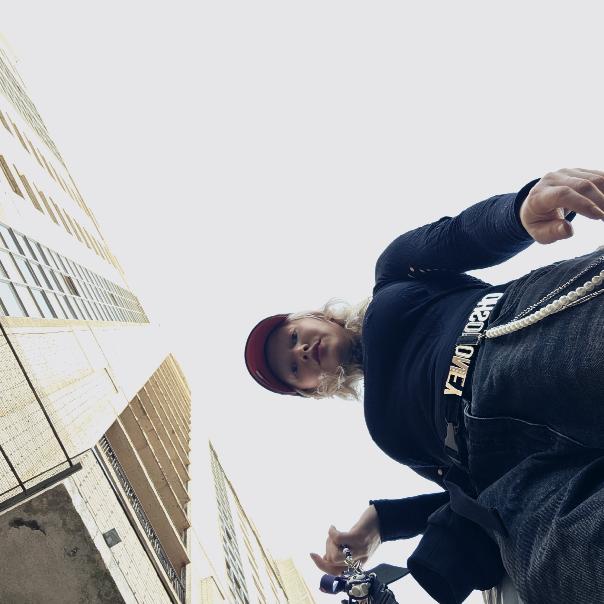 An extreme low-angle photograph captures a light-skinned individual looking directly into the camera from below. The subject wears a red baseball cap, a form-fitting black long-sleeved top, and dark denim jeans. A black belt with a prominent gold-colored buckle, displaying the reversed text "Y3MO 02HO," is visible at their waist. Multiple decorative chains, including a strand of white pearls and several metallic chains, are suspended from the belt. Light blonde, wavy hair frames their face, and a dark tattoo is visible on the left side of their neck. In their raised left hand, they hold a multi-component object, possibly a compact camera or a keyring with a decorative charm. The background features a bright, uniformly white sky, against which two large, light-colored multi-story buildings with numerous visible windows rise vertically and diagonally, suggesting a modern urban environment. The scene is located in the city of Saint Petersburg, Russia.