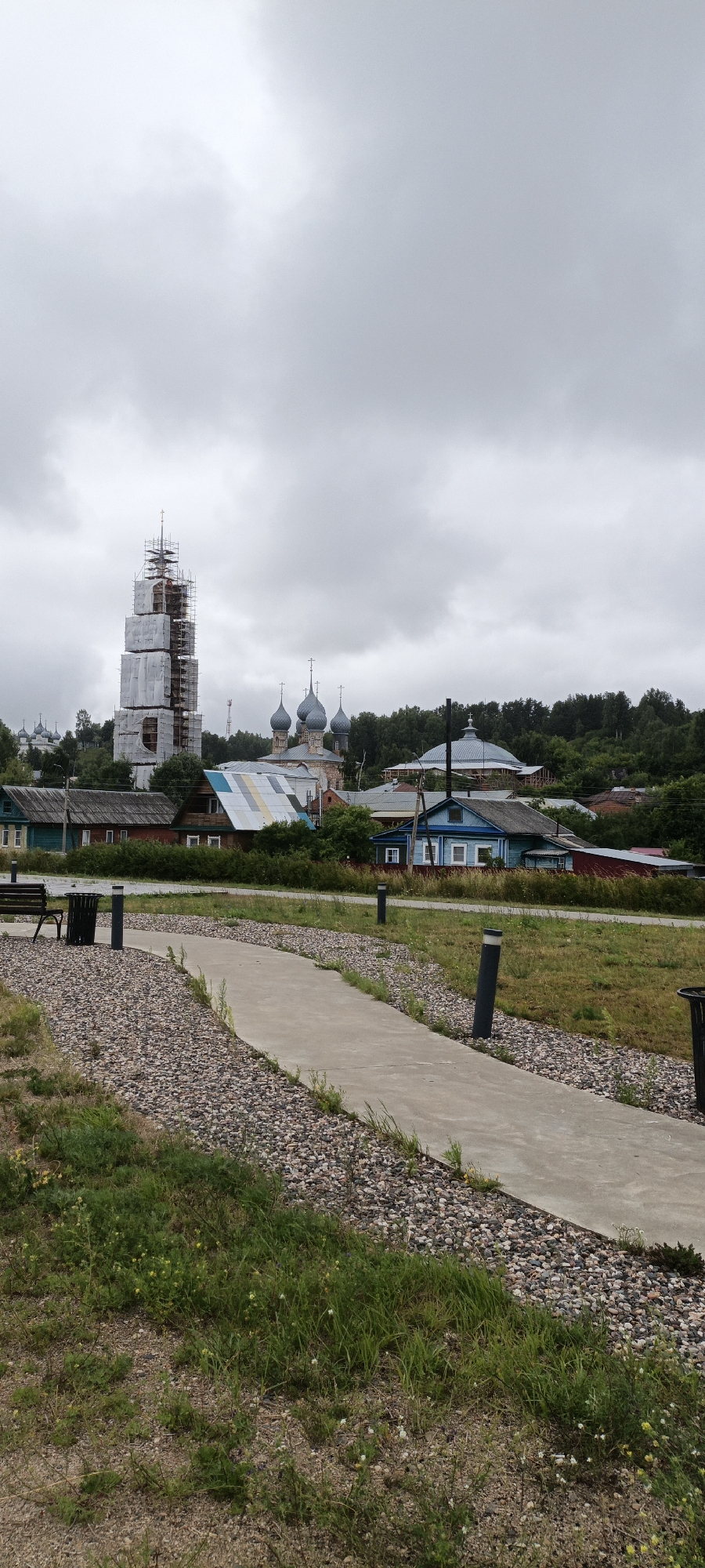 In Yuryevets, Russia, under an overcast sky, a paved pathway meanders through a landscape of mixed gravel and grass. To the left, a dark park bench and a cylindrical trash receptacle sit near the path's beginning. In the mid-ground, a row of village houses, constructed from wood and featuring colorful tin roofs, stretches across the scene. Behind the houses, a complex of church buildings with distinctive blue onion domes stands, and further to the left, a tall, bell tower is partially encased in scaffolding and white sheeting, suggesting ongoing construction or renovation. A dense line of green trees forms a backdrop to the settlement, with a gentle hill visible beyond. The overall impression is one of a rural village with historical architecture, actively undergoing preservation.