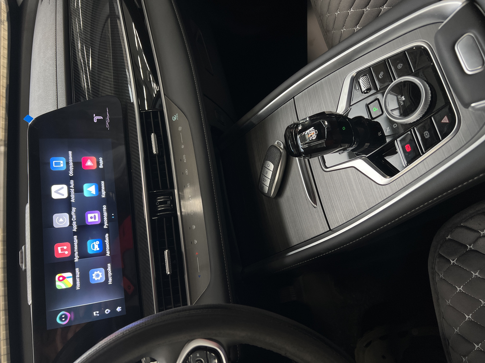This image depicts the interior of a modern vehicle located in Izhevsk, Russia. The scene focuses on the dashboard and center console, presented from the driver's perspective.
Dominating the dashboard is a large, rectangular infotainment touchscreen display. The screen displays a grid of application icons with Russian labels, including "Навигация" (Navigation), "Мультимедиа" (Multimedia), "Apple CarPlay", "Android Auto", "Оборудование" (Equipment), "Настройки" (Settings), "Автомобиль" (Car), "Руководство" (Manual), "Картинки" (Pictures), and "Видео" (Video). A stylized "T" logo above the word "Tiggo" is visible on the screen. A small blue adhesive strip is attached to the upper right edge of the screen bezel. Below the screen, a panel features horizontal air vents and a series of climate control buttons and indicators, including temperature adjustment, fan speed, A/C, "AUTO" mode, and "PURE" air quality mode. The dashboard features a dark, carbon fiber-look trim extending across the passenger side.
The center console, finished with a dark, brushed metallic or wood-grain texture, houses the primary vehicle controls. A black and silver car key fob rests on the console. An electronic gear selector, translucent black with a distinct tiger head logo at its apex, is positioned prominently. Surrounding the gear selector are "P" (Parking), "R" (Reverse), "N" (Neutral), and "D" (Drive) buttons. A large, chrome-trimmed rotary dial labeled "MODE" is centrally located, accompanied by other control buttons for functions such as the electronic parking brake (a red icon with 'P'), auto-hold, hazard lights (a red triangle), and other driving assistance or menu controls. The left side of a multi-function steering wheel, in a dark material, is partially visible on the left edge of the frame. A quilted pattern is visible on the upholstery of both the driver's and passenger's seats, indicating a premium interior finish. The interior appears clean and well-maintained, suggesting the vehicle is new or in a showroom environment.