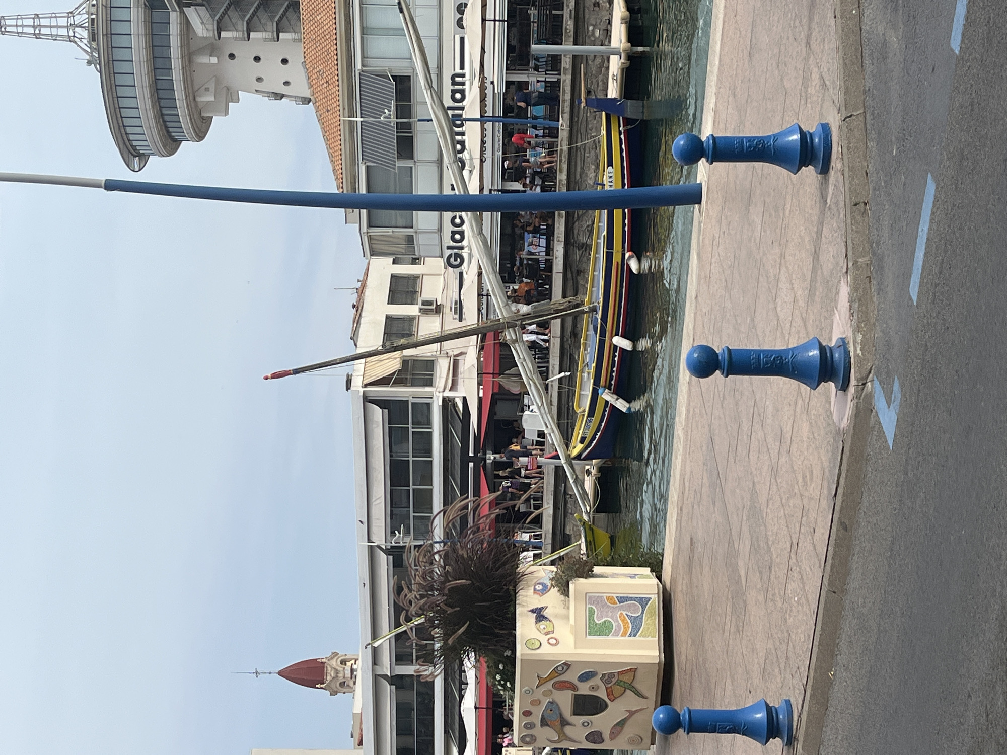 The image depicts a view of the marina in Palavas-les-Flots, France, with several elements of the town visible. In the foreground, a paved walkway lines the harbor, marked by decorative blue bollards. Beyond the walkway, the water of the marina is visible, reflecting the sky and surrounding structures.
Dominating the center of the image is a traditional Mediterranean fishing boat, painted with bright yellow, blue, and red stripes. The boat has a single mast and sail, and it is moored along the waterfront. Next to the boat, a planter filled with ornamental grass sits on the edge of the walkway, decorated with colorful mosaic tiles featuring fish and abstract patterns.
In the background, a row of restaurants and shops line the harbor, with awnings extended over outdoor seating areas where people can be seen dining. Above these buildings, architectural elements of Palavas-les-Flots are visible. To the left, the red-tiled roof and steeple of a church tower pierce the sky. To the right, a distinctive modern observation tower with a circular structure and large windows stands out against the sky. The overall scene is one of a vibrant seaside town, with a blend of traditional maritime elements and modern urban architecture.