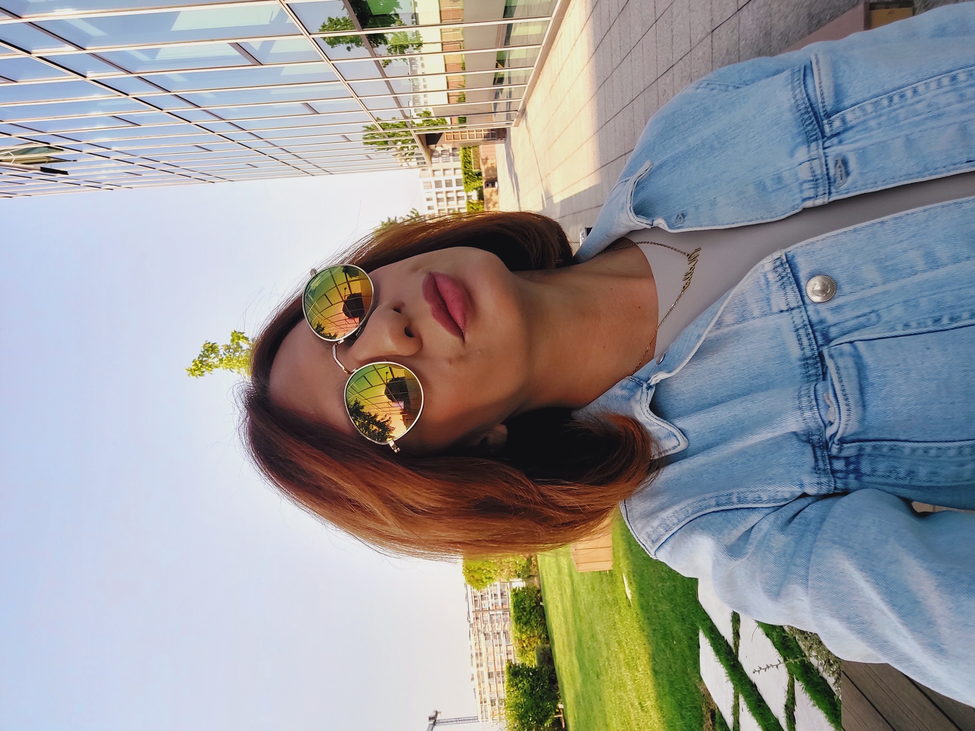 A medium shot selfie of a woman with auburn hair, wearing round, reflective sunglasses and a light blue denim jacket over a gray t-shirt, is captured outdoors. She is looking up and slightly to the right, with her lips pursed. Her necklace with gold lettering is visible. The background features a modern glass building with reflective windows, a manicured lawn, and a construction crane in the distance, suggesting an urban setting. The lighting indicates it is daytime, likely late afternoon due to the warm reflections in her sunglasses and the long shadows. The setting is consistent with the area around the National Palace of Culture in Sofia, Bulgaria, known for its contemporary architecture and green spaces.