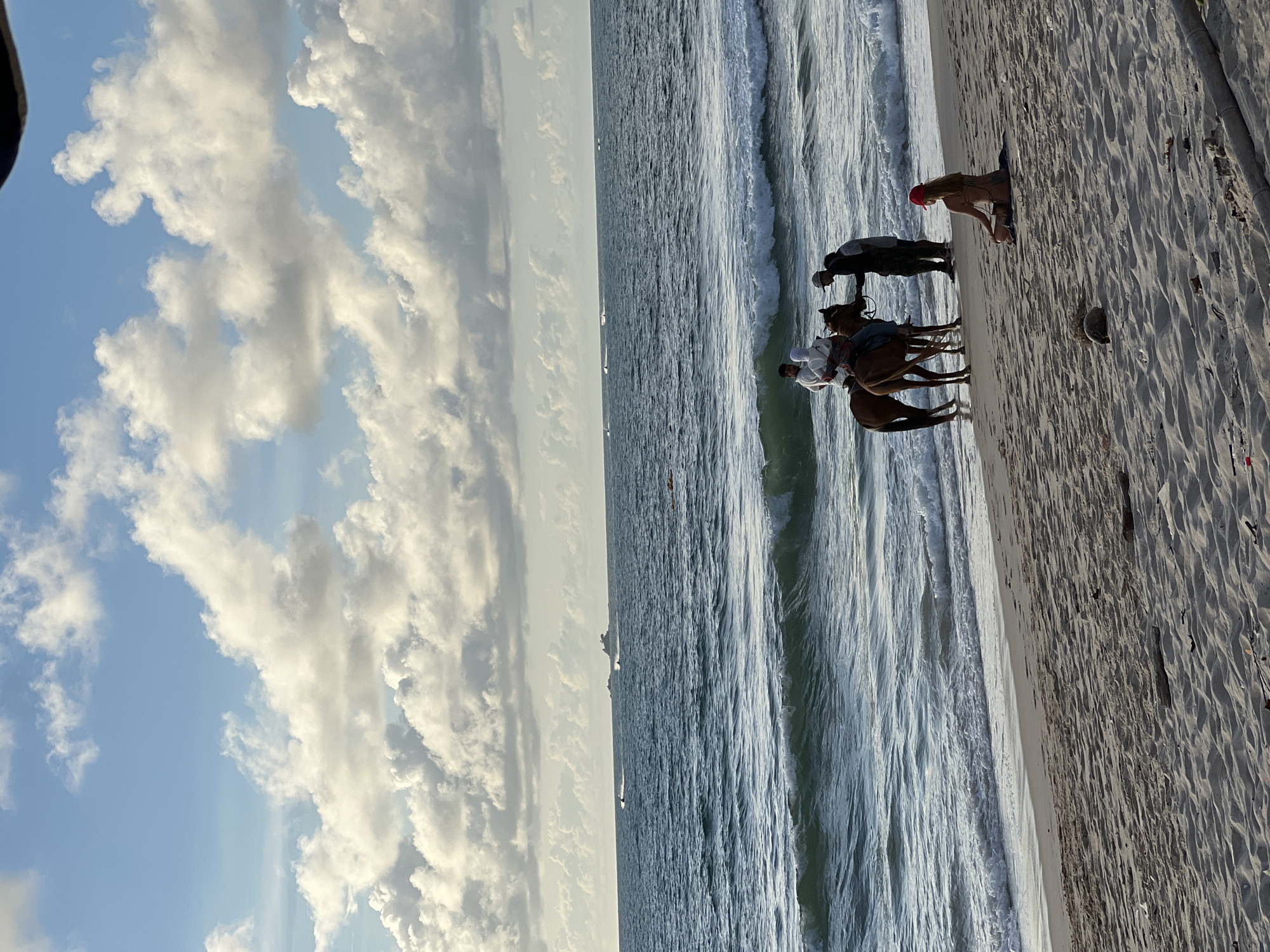 On a sandy beach in Choeng Thale, Thailand, two people are interacting with a horse and rider near the water's edge. The rider is on a brown horse, and two individuals stand next to the horse, one appearing to be talking to the rider. In the foreground, a person wearing a red cap sits on the sand, facing the ocean. The ocean has gentle waves, and in the distance, several jet skis are visible on the water, along with a larger vessel on the horizon. The sky is partly cloudy with a mix of blue sky and white clouds.