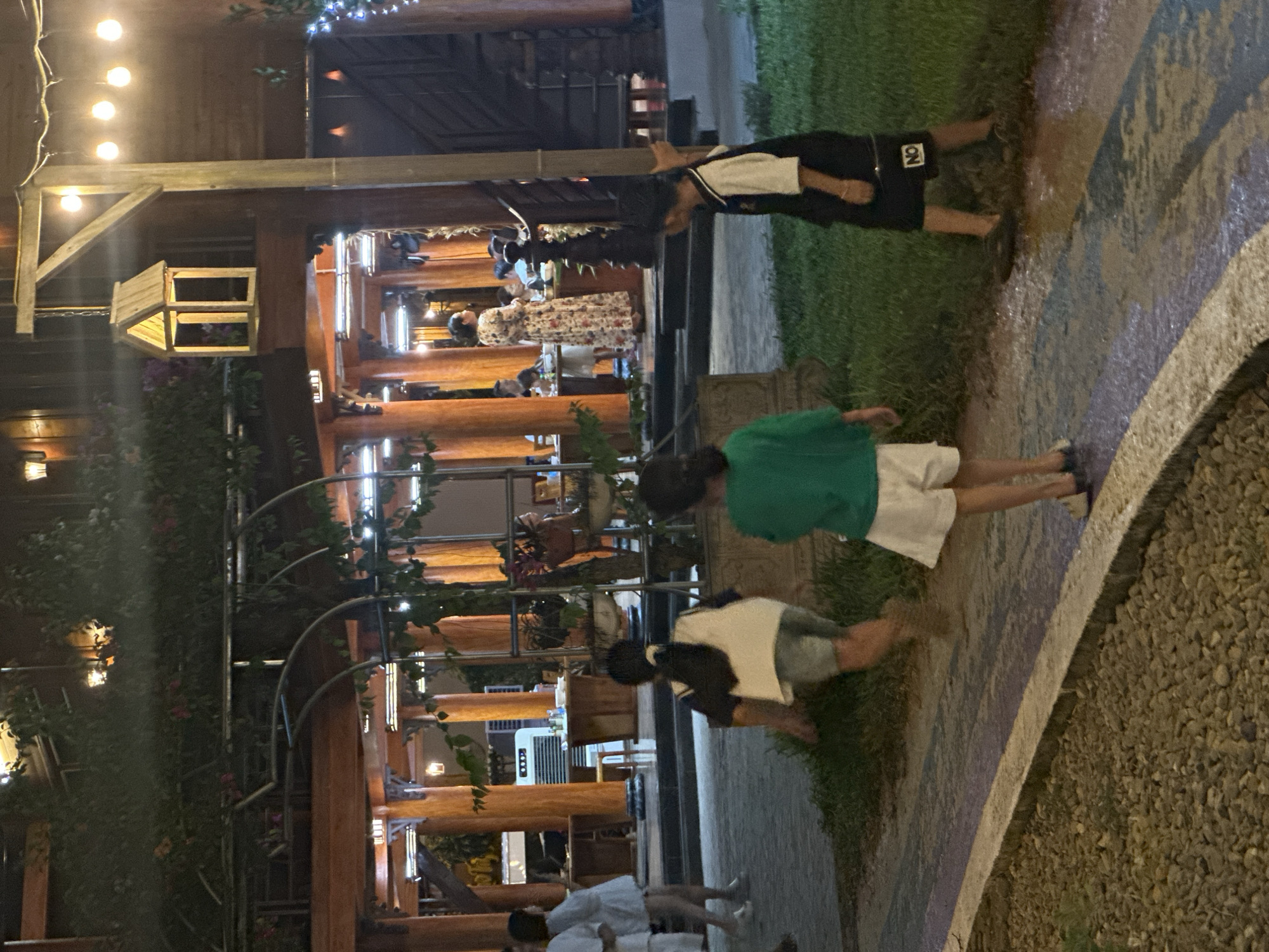 The image depicts a nighttime outdoor scene in Nghia Lo Ward, Vietnam, centered around a large, open-air wooden structure that appears to be a restaurant or community building. The structure features numerous prominent wooden pillars and beams, with some sections covered by climbing plants or vines. Various light sources illuminate the scene, including warm-toned string lights suspended overhead, wall-mounted lanterns, and brighter internal lighting within the building. A decorative wooden lantern, shaped like a miniature house, hangs from a wooden post on the right side of the frame.
In the foreground, a paved pathway with a concrete curb separates a gravel area on the left from a grassy patch on the right. A wet or muddy area is visible on the grassy patch and partially on the pathway, reflecting some of the artificial light. Three children are present in the immediate foreground and midground, appearing to move or play near this wet area. One child, seen from behind on the left-center, wears a dark top with a white panel and light shorts, with legs blurred indicating motion. Another child, also seen from behind in the center, wears a green long-sleeved shirt and white shorts, standing still. A third child on the right, partially obscured by a pole, wears a black and white shirt and black shorts with "NO" printed on one leg, standing in the wet ground.
In the midground, to the left of the main building, two individuals are walking away from the structure. One wears a white shirt and white shorts, while the other is dressed in a light-colored top or dress with white shoes. Inside the brightly lit wooden building, multiple people are seated at tables, suggesting dining or socializing. A woman in a long floral dress stands centrally within the building, facing left, appearing to interact with or supervise guests. An air conditioning unit or large fan is visible inside the building on the left. On the far right, a dark, vertical structure, possibly a gate or wall, is partially visible. The overall lighting suggests an evening setting, with artificial illumination creating focal points and casting shadows.