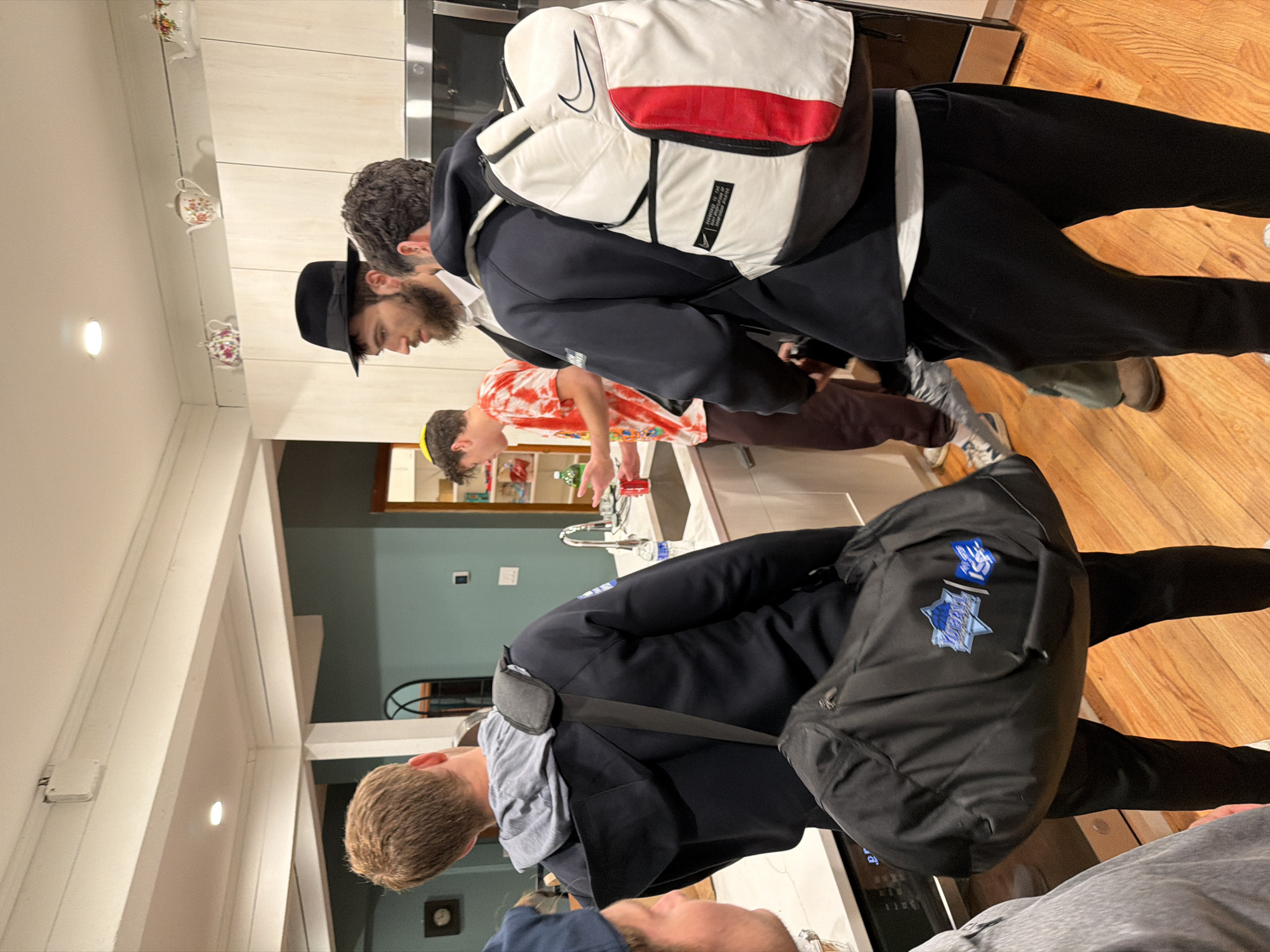 The image shows several people indoors, possibly in a kitchen setting. One man is wearing a black hat, a navy blazer, and carrying a white and red backpack. Another man is wearing a red and orange tie-dye shirt. A third person is wearing a navy jacket over a grey hoodie, and a large black backpack. There are white cabinets and wooden floors. There is no definitive evidence within the image to confirm its location in Rock Hill, United States of America.