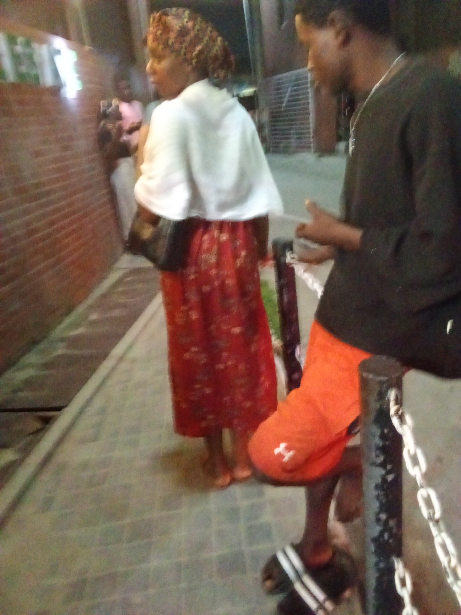 The image shows a street scene in Ikorodu, Nigeria. A woman stands with her back to the camera, wearing a red floral print skirt, a white shawl, and a head covering. She is looking towards a group of people near a brick wall. A man in a black shirt and orange shorts is sitting on a metal post with a chain; his right leg is visible. He is looking down at his hands. The ground is paved with bricks.