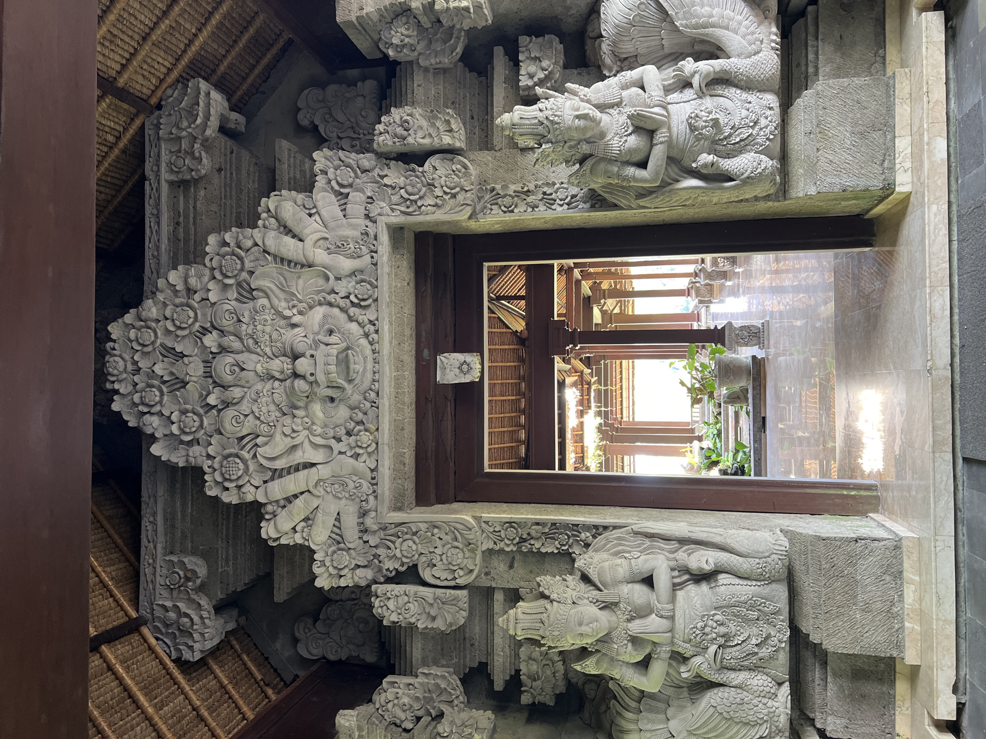 The image captures an ornately carved stone doorway leading into an interior space. The doorway's lintel and surrounding structure are densely decorated with floral patterns, stylized figures, and mythical creatures. The carvings exhibit a high level of detail and craftsmanship. Through the doorway, the interior reveals a colonnaded walkway with a tiled floor, plants, and wooden architectural elements. The location is Kedewatan, Indonesia, as it is known for its traditional Balinese architecture and stone carvings.