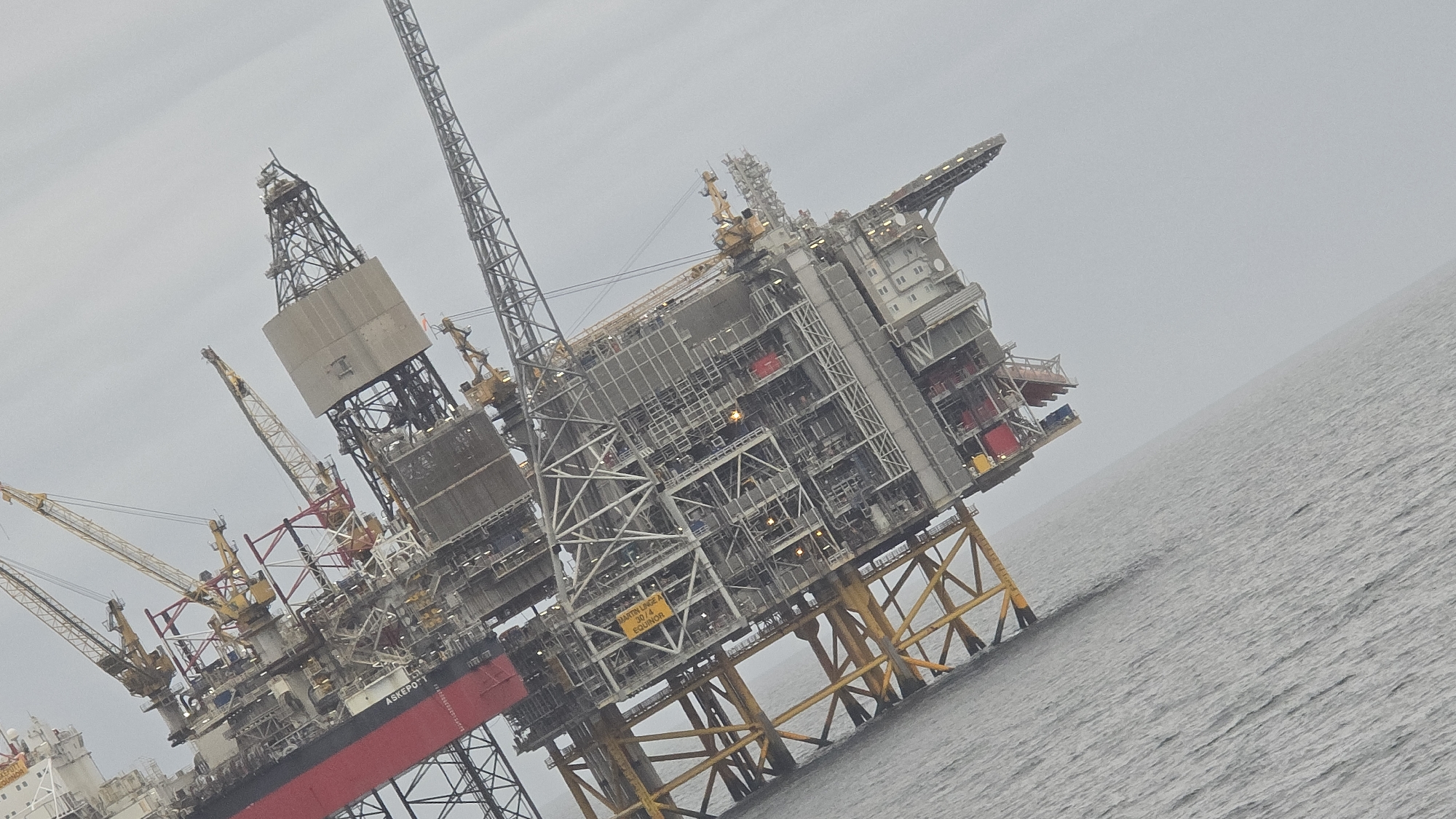 The image shows an offshore oil platform at sea. The platform has multiple structures, including drilling derricks, cranes, and accommodation modules. The platform appears to be standing on steel legs, which are submerged in the water. The sky is overcast, suggesting possibly poor weather conditions. The platform's name ASKEPOTT is visible in the bottom left corner. A yellow sign reads "MARTIN LINGE A 30/4 EQUINOR" can also be seen on the platform. There is a ship docked on the side of the oil platform.