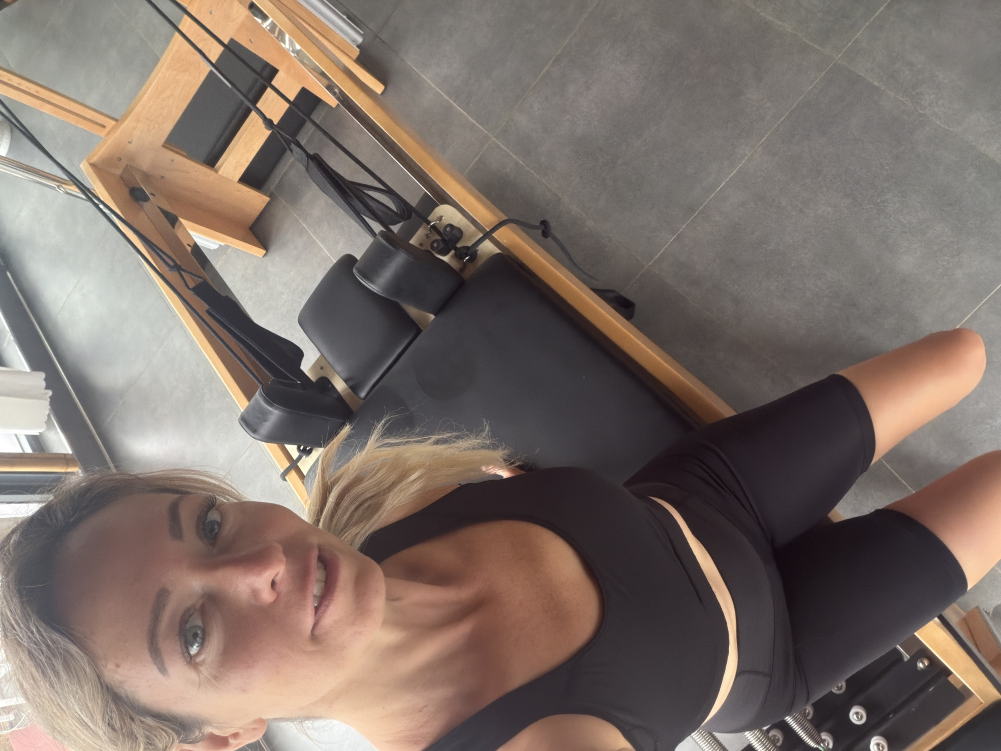 The image shows a woman with blonde hair, wearing a black sports bra and black shorts, sitting on a Pilates reformer. She is looking at the camera with a slight smile. The Pilates reformer is made of wood and has black padding. The floor is tiled with large gray tiles. The scene appears to be indoors, possibly in a gym or studio. There are no discernible landmarks or features that would indicate the specific location within Da Lat, Vietnam.