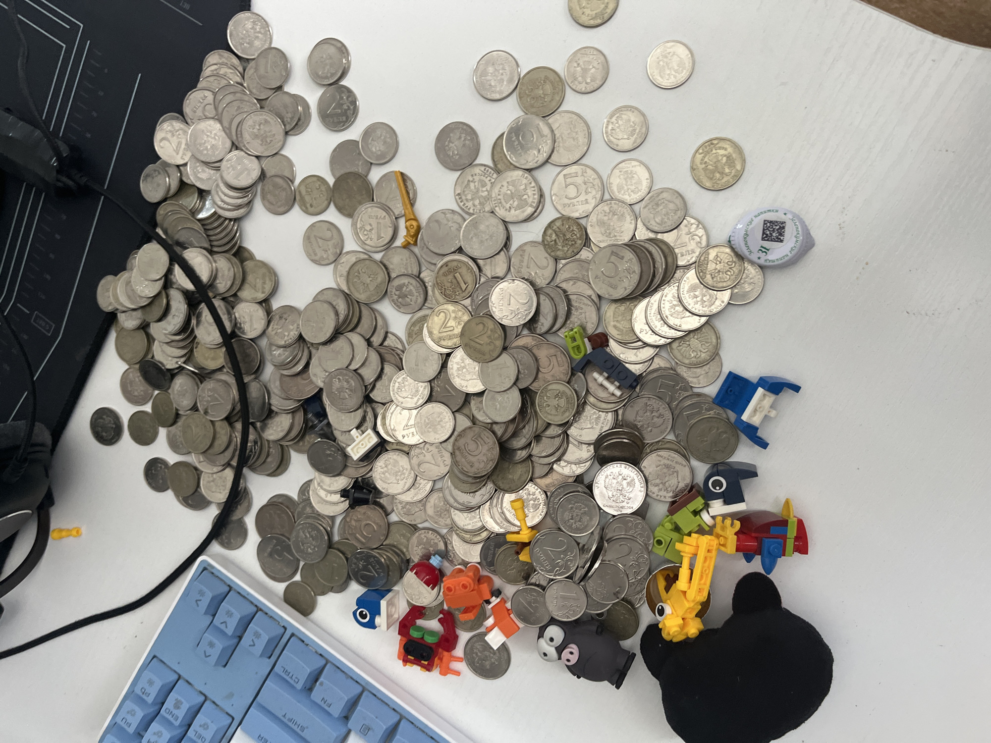 A close-up, high-angle shot captures a white desk surface littered with numerous silver-colored coins, some stacked and others scattered. Interspersed among the coins are several small, colorful plastic building toy pieces, including a red, orange, and yellow structure, a blue and white piece, and a green and blue character with a red body. A grey pig figurine with wide eyes and a black plush toy resembling a bear are also present on the desk. A portion of a light blue mechanical keyboard is visible in the upper left corner, and a black gaming mousepad occupies the upper right. A black cable snakes across the desk, connecting to unseen devices. The overall scene depicts a collection of everyday objects on a workspace. The location is not specified in the image, and therefore cannot be determined to be Saratov, Russia.