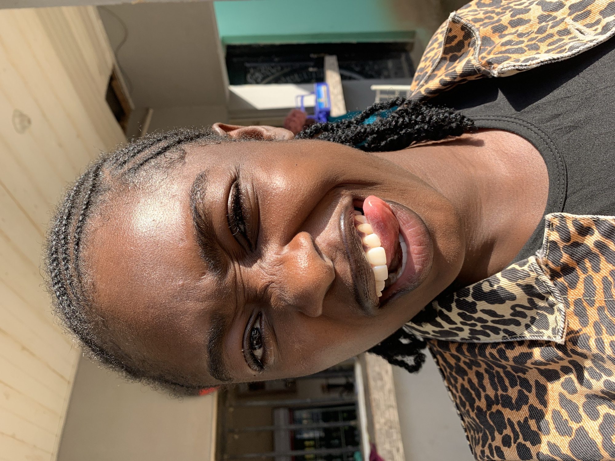 A dark-skinned individual is presented in a close-up, head-and-shoulders portrait in New Stadium, Nigeria. The individual exhibits a playful facial expression, characterized by squinted eyes, a crinkled nose, and an open mouth with the tongue partially extended, touching the upper lip. Braided hair, including cornrows on the scalp and individual braids, is visible, with some blue strands integrated into the braids on the right side. The individual wears a leopard-print jacket over a black round-neck shirt. Strong natural sunlight illuminates the scene, casting highlights on the skin. In the background to the left, a metal security gate or bars are visible through an opening, with indistinct objects behind them. To the right, the background comprises a light-colored wall, a dark doorway or gate, and a section of a teal-colored wall. A light-colored surface with a blue container is present further back on the right. Overhead, a wooden plank ceiling is visible, featuring a circular fixture and an irregular opening revealing a darker cavity.