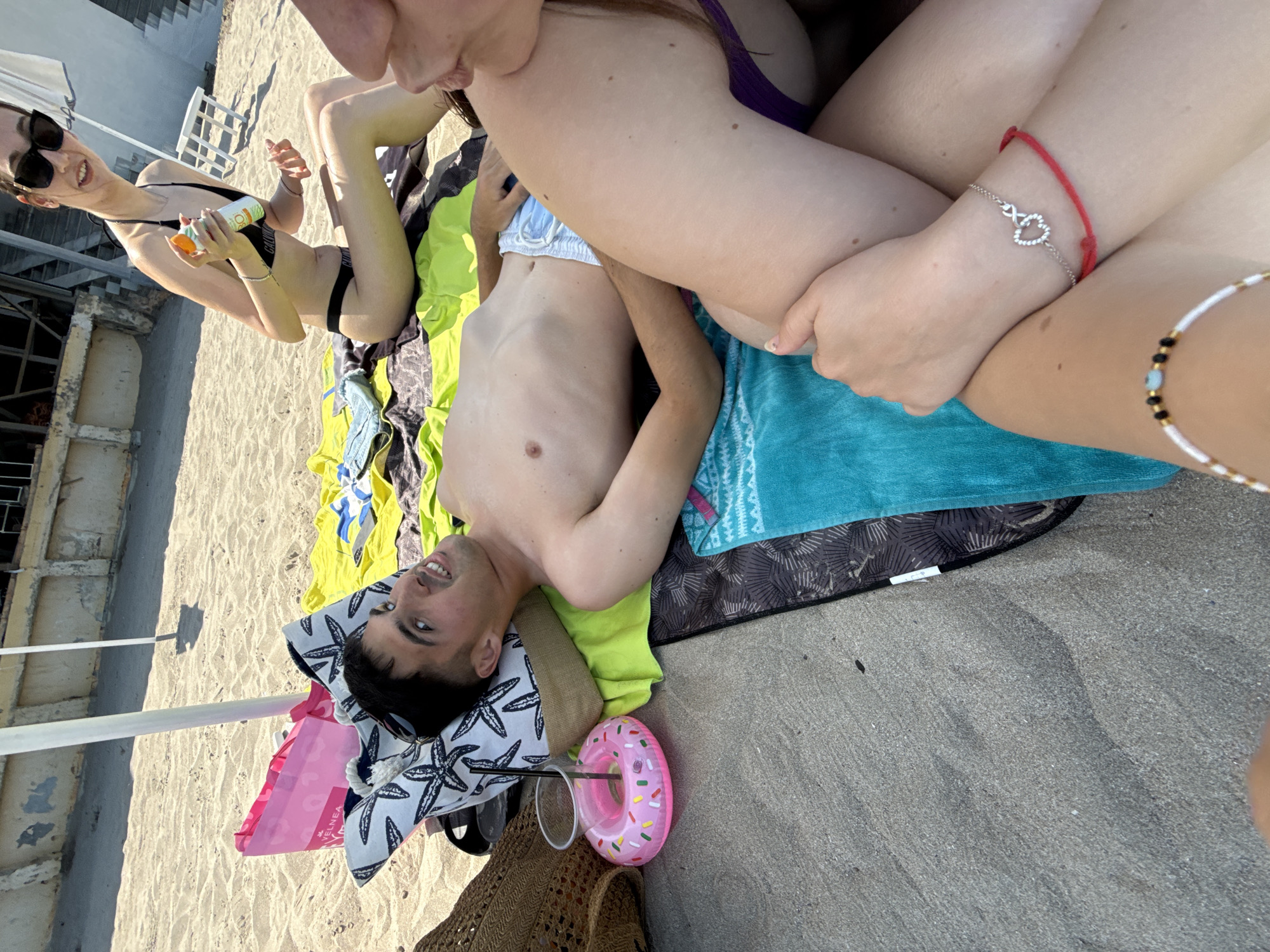 This image captures a relaxed beach scene in Sozopol, Bulgaria, featuring at least three individuals enjoying a sunny day. The shot is taken from a slightly elevated, diagonal perspective, looking down at the group on the sand.
In the mid-left of the frame, a young woman with dark hair, wearing sunglasses and a black bikini with a visible "Calvin Klein" band, smiles towards the camera. She is holding a bottle of sunscreen or lotion, appearing to be applying it or preparing to. Her legs are bent, and she is partially reclined on a beach towel.
Diagonally below, a young man lies on his back, shirtless, resting his head on a lime green towel and a pillow or towel with a starfish pattern. He looks up with a pleasant smile. Next to him is a pink, donut-shaped floating drink holder with a clear cup and straw. A partially visible woven straw bag and a pink bag, which bears the text "YELNEA," are also nearby.
To the right, the lower body and arms of another person are visible. This individual wears a red string bracelet and a silver bracelet adorned with an infinity symbol and a heart on one wrist, and a beaded bracelet on the other. A glimpse of a purple bikini can also be seen. This person appears to be situated closely with the man and the other woman.
The setting is a sandy beach, with strong sunlight indicating a bright, clear day. In the background, there is a light-colored building with stairs, possibly a beach facility. The overall atmosphere suggests a leisurely and enjoyable vacation or outing.
