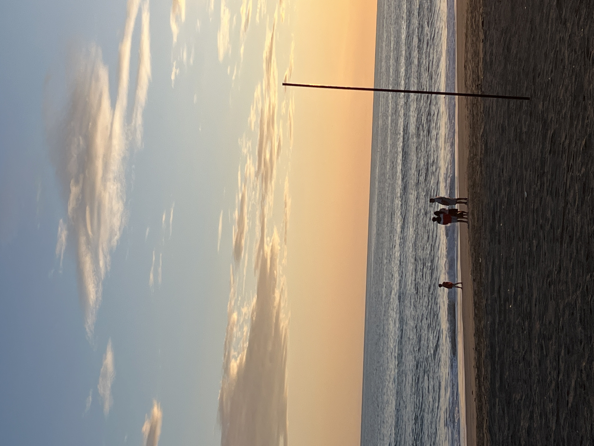 Here is a description of the image:
The image captures a serene beach scene at sunset. The sky transitions from blue with scattered clouds on the left to a warm orange hue towards the center. A body of water, likely the sea, occupies the central portion of the image with gentle waves visible. A dark, sandy beach stretches along the right side of the frame.
Silhouetted figures of several people are present on the beach. A thin, dark line runs horizontally across the image, potentially a wire or rod.
Based on the description and the "Alanya, Turkey" context, it is likely that the beach is located in or near Alanya, a coastal city known for its beaches. However, without a specific landmark, pinpointing the exact location within the city is not possible.