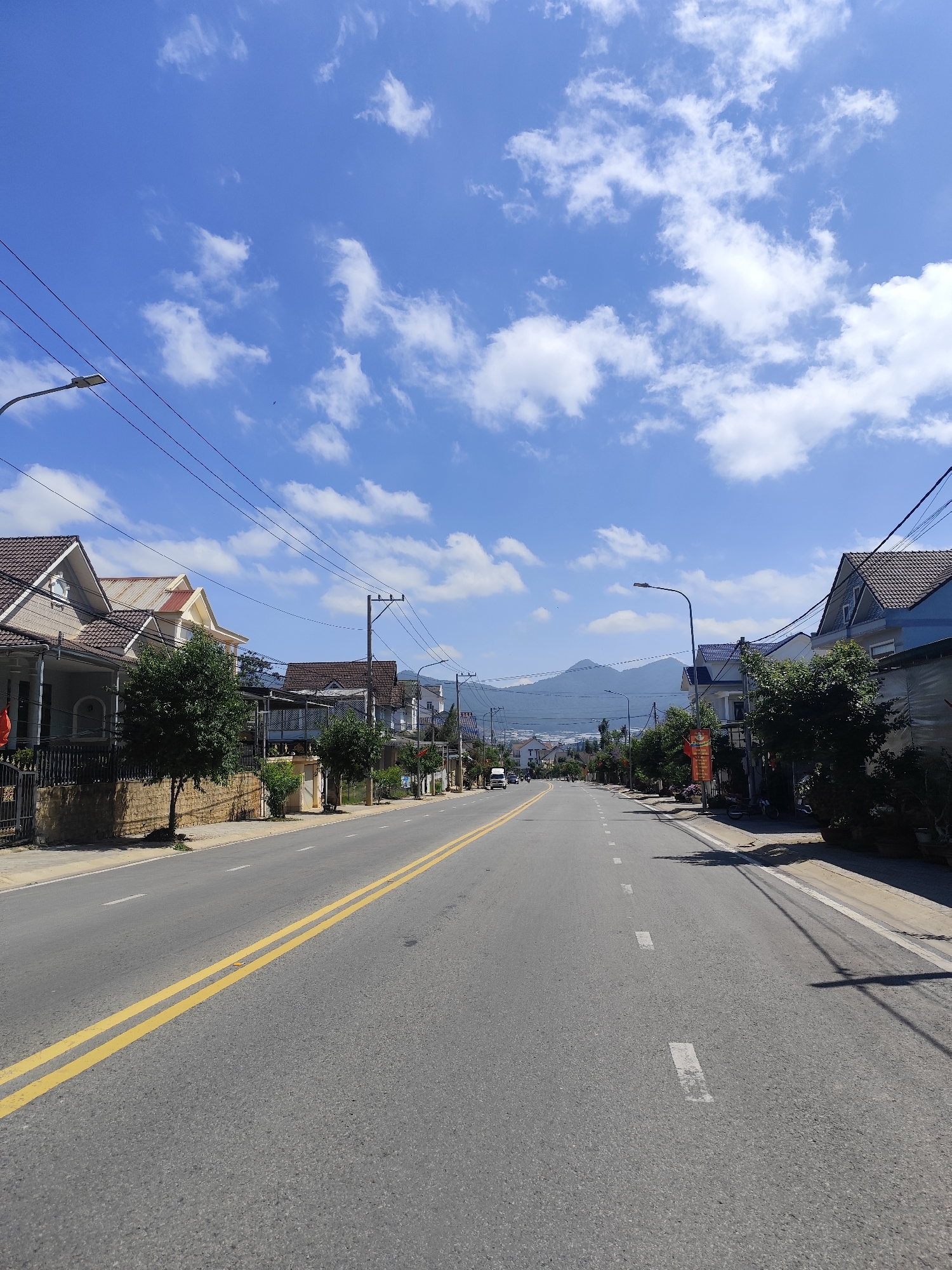 This vertical image depicts a peaceful, wide asphalt road stretching into the distance under a bright blue sky dotted with scattered white clouds. The scene is a residential street, likely in Lạc Dương, Vietnam, with distant mountains forming a hazy backdrop.
The road features clear yellow double lines marking the center, and white dashed lines on the right side. Both sides of the street are lined with modern, predominantly two-story houses, each with pitched roofs varying in color from brown and gray to blue. Small trees and shrubs are planted along the sidewalks in front of the homes, often separated by low stone walls or metal fences. Overhead, numerous utility poles support a dense network of electrical wires, and streetlights are visible along the route.
The time of day is clearly daytime, evidenced by the bright sunlight casting distinct shadows from the utility poles and trees. The weather is sunny and clear, contributing to the serene atmosphere. No people are visible on the street, and only a few indistinct vehicles can be seen far down the left lane, suggesting a quiet moment. A red banner or sign with unreadable white text is visible near a house on the right, and a small red flag is partially seen on the left. The overall impression is one of calm and tranquility in a suburban setting.