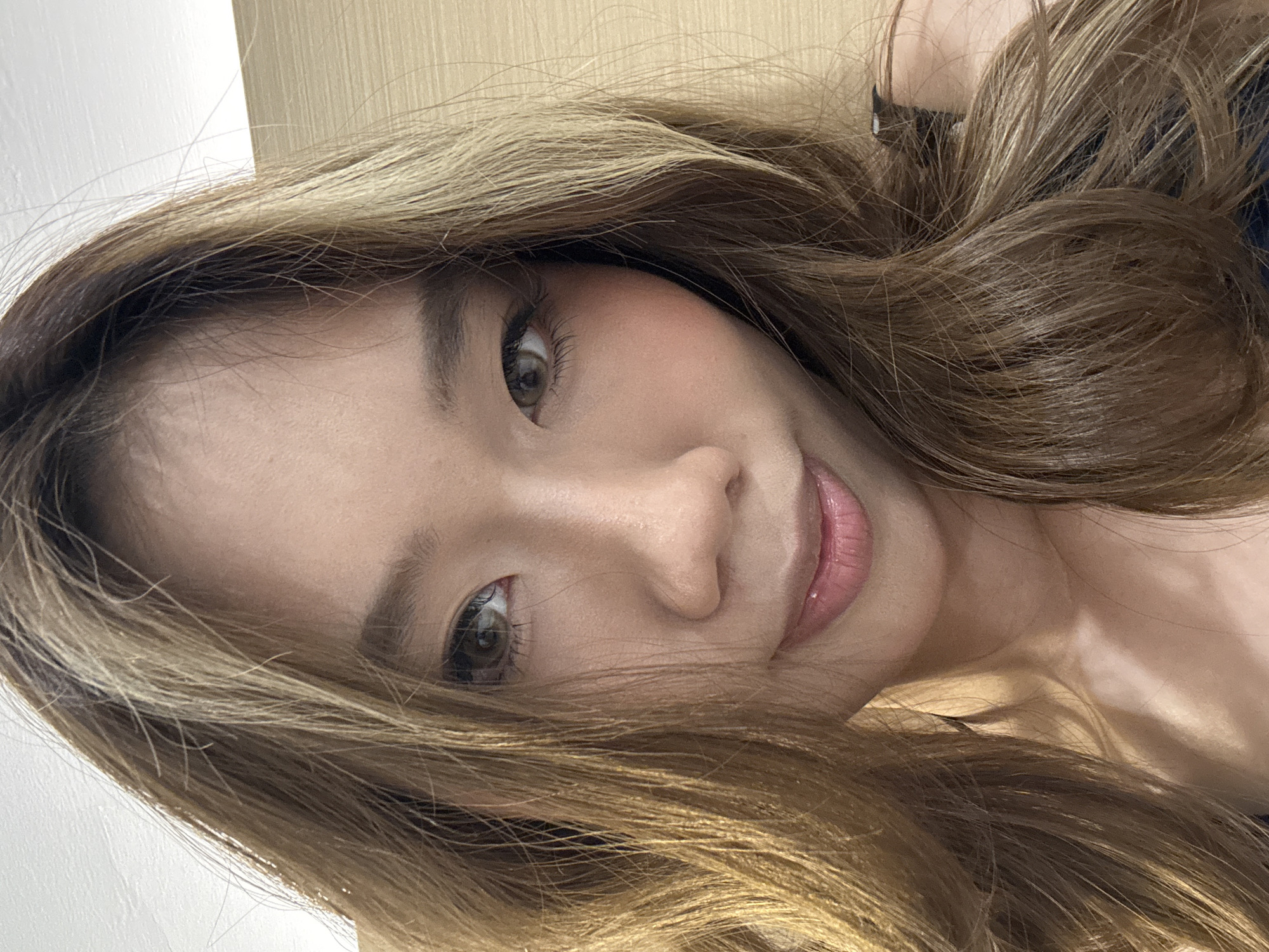 A close-up portrait shows a young woman with long, wavy, light brown hair, interspersed with lighter, caramel-blonde highlights. Her head is tilted, and her hair cascades around her face and shoulders, filling much of the frame. She has a fair complexion with a hint of rosy blush on her cheeks. Her large, hazel or dark brown eyes gaze directly at the viewer, framed by defined eyebrows and long eyelashes. Her lips are slightly parted in a gentle smile, enhanced with a glossy pink or nude lipstick.
The lighting is soft and even, suggesting an indoor setting. The background is out of focus, revealing a light-colored, possibly white wall or surface on the left, and a textured, light brown surface, perhaps wood paneling, on the right. A glimpse of dark clothing, possibly a strap or top, is visible beneath her hair on the right side. The overall expression is calm and pleasant.
The activity taking place is a close-up selfie or portrait. There is no visible text in the image. The location is given as Yuzhong District, China, though no specific visual cues confirm this.