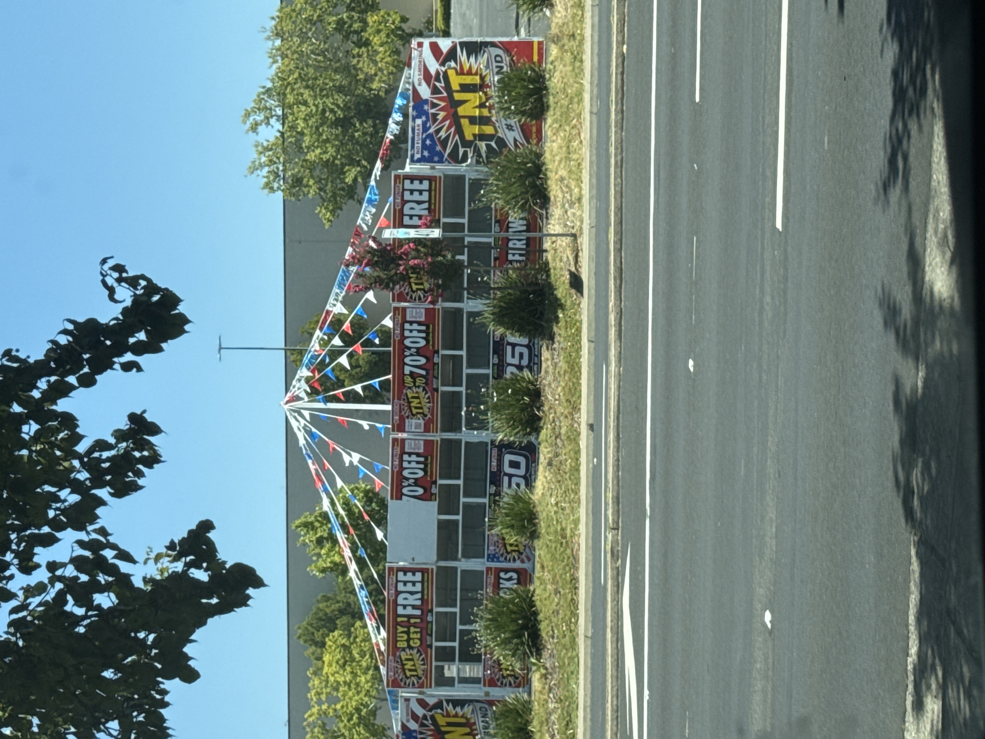 A temporary fireworks stand is set up in a commercial area of Citrus Heights, California, USA, likely in a parking lot. The stand has a white roof supported by poles, adorned with red, white, and blue triangular flags. The front of the stand features multiple signs advertising sales and promotions for "TNT" brand fireworks. Some visible signs include "BUY 1 GET 1 FREE", "70% OFF", and "UP TO 70% OFF". Below these signs, there are windows revealing rows of fireworks. The stand is surrounded by green bushes and a grassy area. In front of the stand is a multi-lane road with white lane markings. The background shows a building and trees. The sky is clear and blue, and sunlight casts shadows.