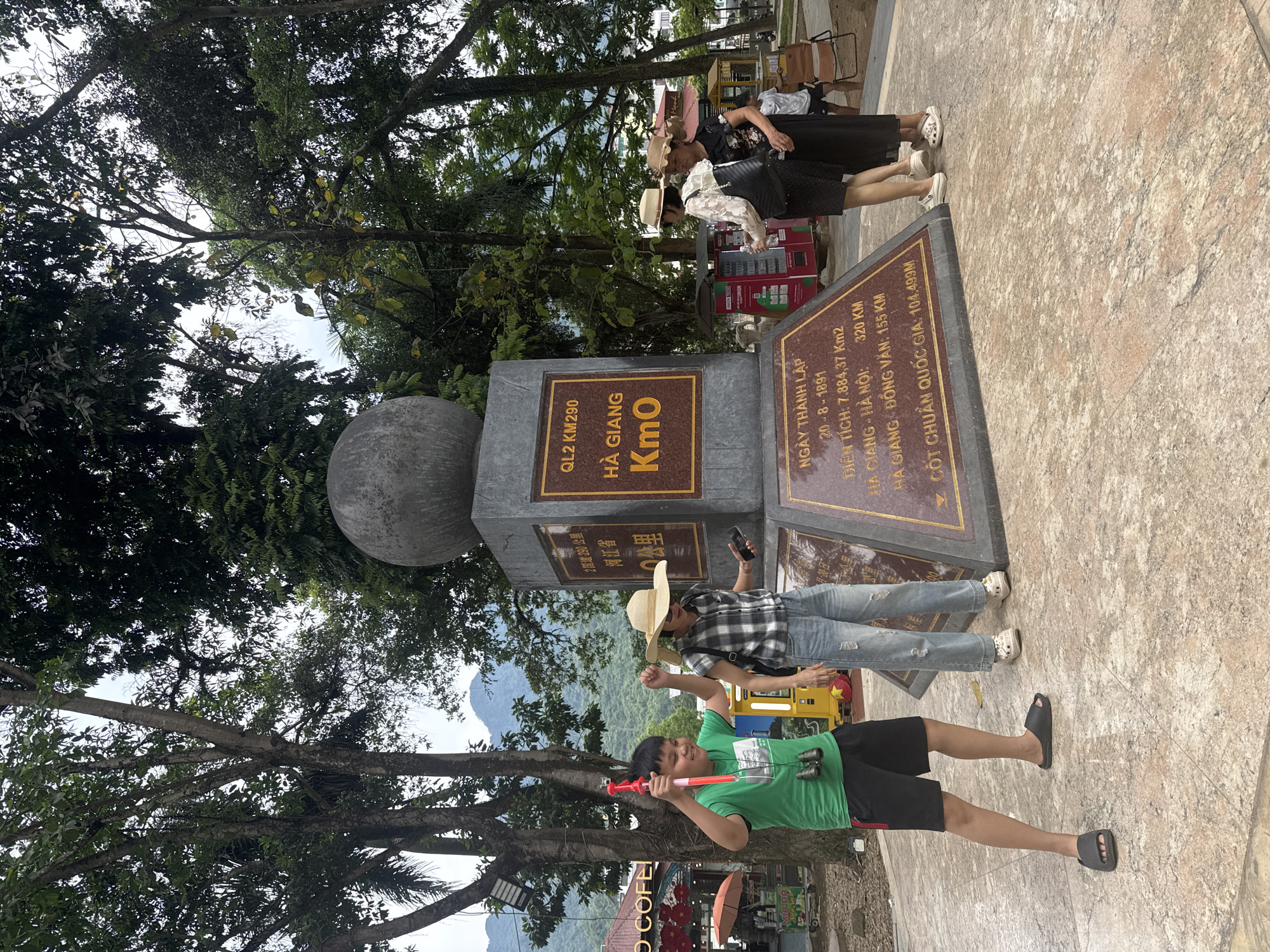 Here's a concise description of the image:
The image shows a group of people near a Km0 landmark in Hà Giang 1 Ward, Vietnam. The landmark features a stone pillar with a spherical top and a plaque with details about the province. Two women stand on the landmark at the top edge of the image with light-colored sun hats. A man in a plaid shirt and a large sunhat holds a smartphone, posing near the landmark. Below the man stands a child wearing a green t-shirt and a pair of binoculars, holding a red and pink toy. Trees surround the landmark, and mountains are visible in the background.