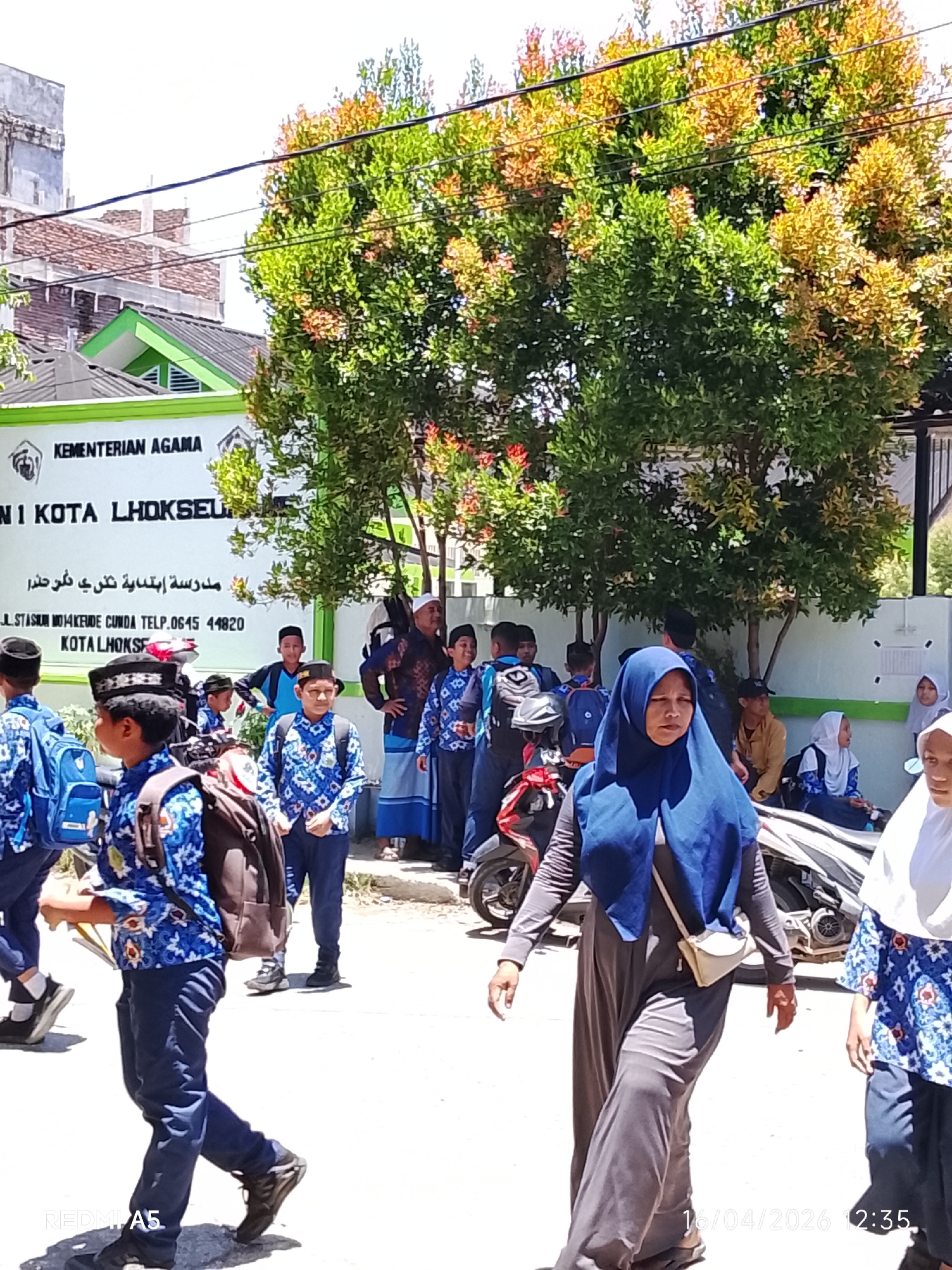 The image shows a group of students in blue and white uniforms outside a building that reads "KEMENTERIAN AGAMA NI KOTA LHOKSEU." The text below the sign in Arabic reads "Madrasah Ibtidaiyah Negeri Fokerde," followed by "JL.STASIUM MO14 KEUDE CUNDA TELP.0645 44820 KOTA LHOKSE." Some students are walking, while others are standing or sitting near a parked motorcycle. An adult woman in a blue hijab and gray clothing is walking in the foreground. The environment appears to be outdoors, with trees in the background and buildings across the street. A date stamp in the bottom right corner shows "16/04/2026 12:35."