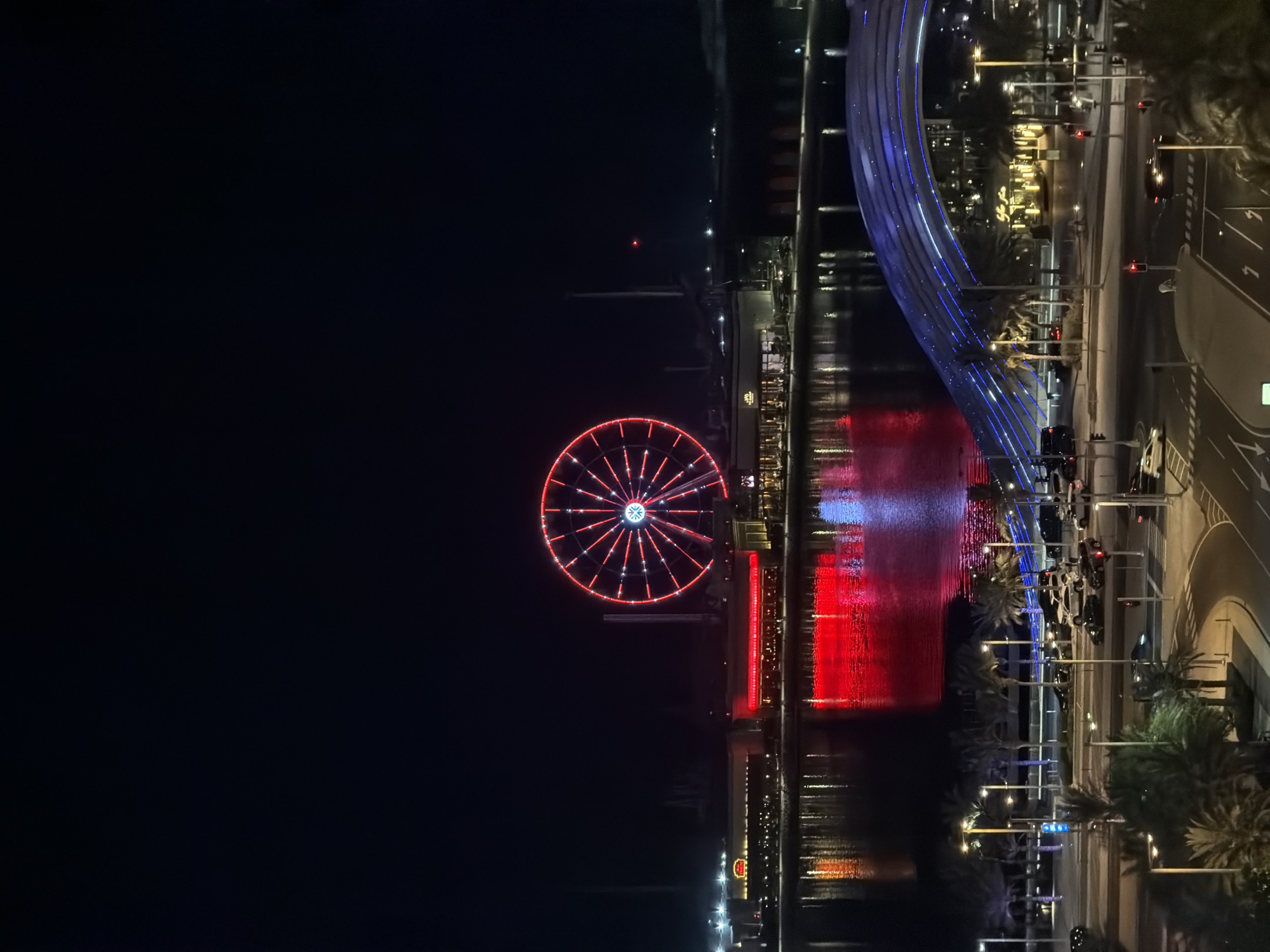 The image depicts a nighttime scene in an urban waterfront area. A large Ferris wheel, illuminated with red lights, is the prominent feature in the background, situated behind buildings lining the water. The buildings are also lit, with red lights reflecting on the water's surface. In the middle ground, a modern bridge with blue lights arches over a road. The foreground shows a street with several cars, palm trees, and streetlights. The sky is dark, indicating it is nighttime. No people are clearly visible in the scene. The overall impression is of a lively, illuminated city environment at night.