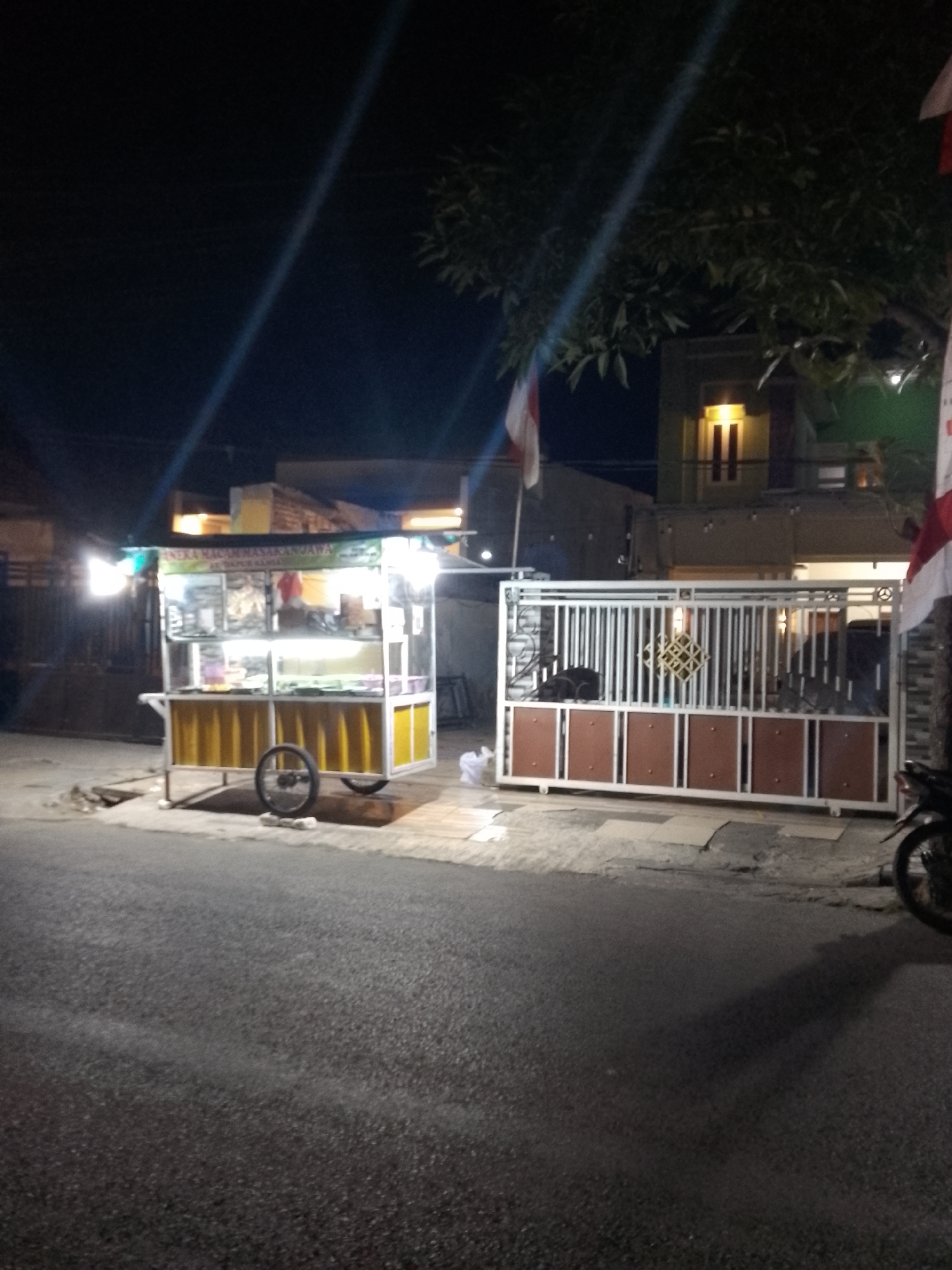 At night in Lamongan, Indonesia, a brightly lit food cart with the words "ANEKA MACAM MASAKAN JAWA" and "ASLI DAPUR BAMBA" is parked on the side of a paved road. The cart is yellow and white, with a transparent display case showcasing food items. A single black bicycle wheel is attached to the cart. Behind the cart is a modern metal gate with brown panels and a decorative diamond pattern, leading to a building with illuminated windows. In front of the gate, a black motorcycle is partially visible on the right side of the image. Indonesian flags are displayed on poles, one to the left of the building and another to the right. The scene is illuminated by artificial lights, creating strong shadows on the road.