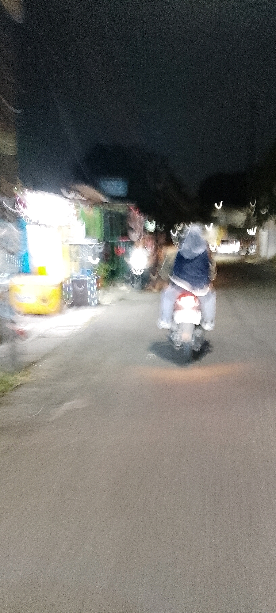 An out-of-focus nighttime photograph captures a person riding a scooter down a street in Bekasi, Indonesia. The rider, wearing a hooded jacket, is the primary subject, their form blurred by motion. To the left, a brightly lit street food cart with a yellow cooler is visible, also affected by motion blur. The background shows indistinct shapes of trees and buildings, with streaks of light suggesting other moving vehicles or streetlights. The pavement is wet, reflecting the ambient light.