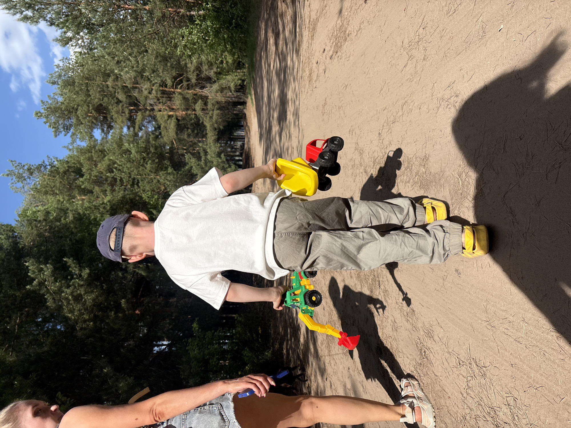 A photograph taken outdoors in the city of Chaikovsky, Russia, shows a person standing on a dirt path, surrounded by trees. The person, a child, is wearing a white t-shirt, grey pants, and yellow shoes. The child is holding two toy vehicles: a yellow and red truck and a green and red excavator. Another person, presumably an adult, is visible from the waist down, wearing denim shorts and sandals. They are holding a blue object in their hand. The ground is mostly dirt with a few scattered blades of grass. Shadows from the surrounding trees and people are cast onto the path.
