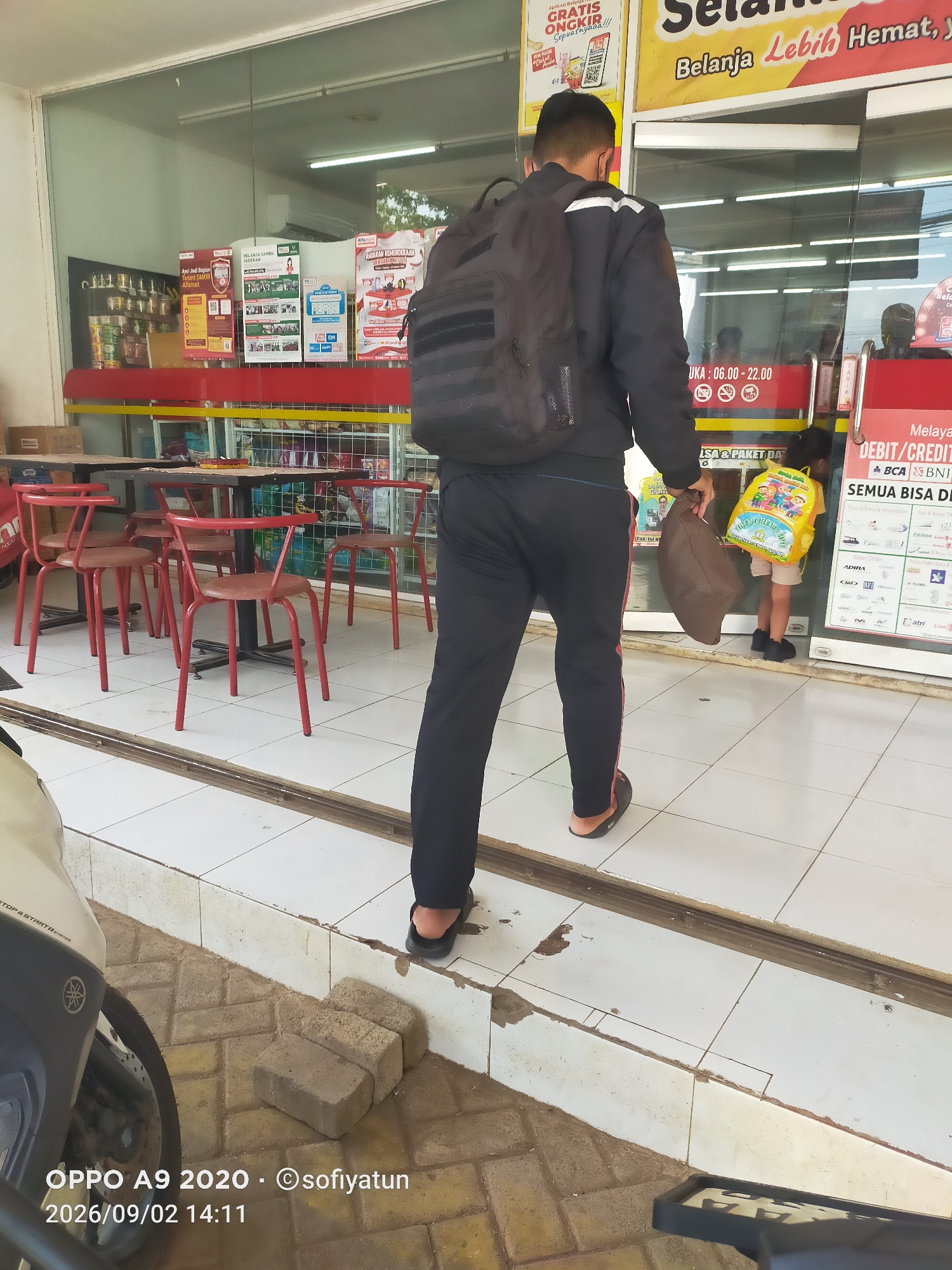 A man wearing a black jacket, black pants with red stripes, and black sandals walks away from the camera, carrying a brown bag in his right hand and a black backpack on his back. He is walking on a tiled pathway in front of a store entrance. Inside the store, a young child wearing a yellow backpack is visible. The store appears to be an Alfamart, a convenience store chain in Indonesia, indicated by the signage and product displays visible through the glass doors. Red chairs and tables are set up outside the store. The timestamp at the bottom left corner indicates the photo was taken on September 2, 2026, at 2:11 PM, and was taken with an OPPO A9 2020. The location is identified as Tayu, Indonesia, consistent with the presence of an Alfamart.