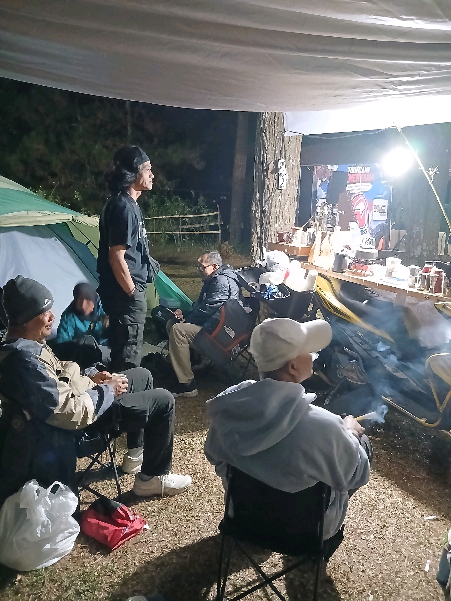 A group of people are gathered around a campfire at night in an outdoor setting, likely a campsite. The scene appears to be in the village of Berjo, Indonesia. Several individuals are seated in camping chairs, some holding cups. One person stands, looking away from the camera, while others are partially visible in the background. Tents are set up, and a makeshift table displays various items, possibly for a coffee station, with a bright light illuminating the area. Smoke from the campfire rises into the air. The ground is covered in grass and scattered wood chips.