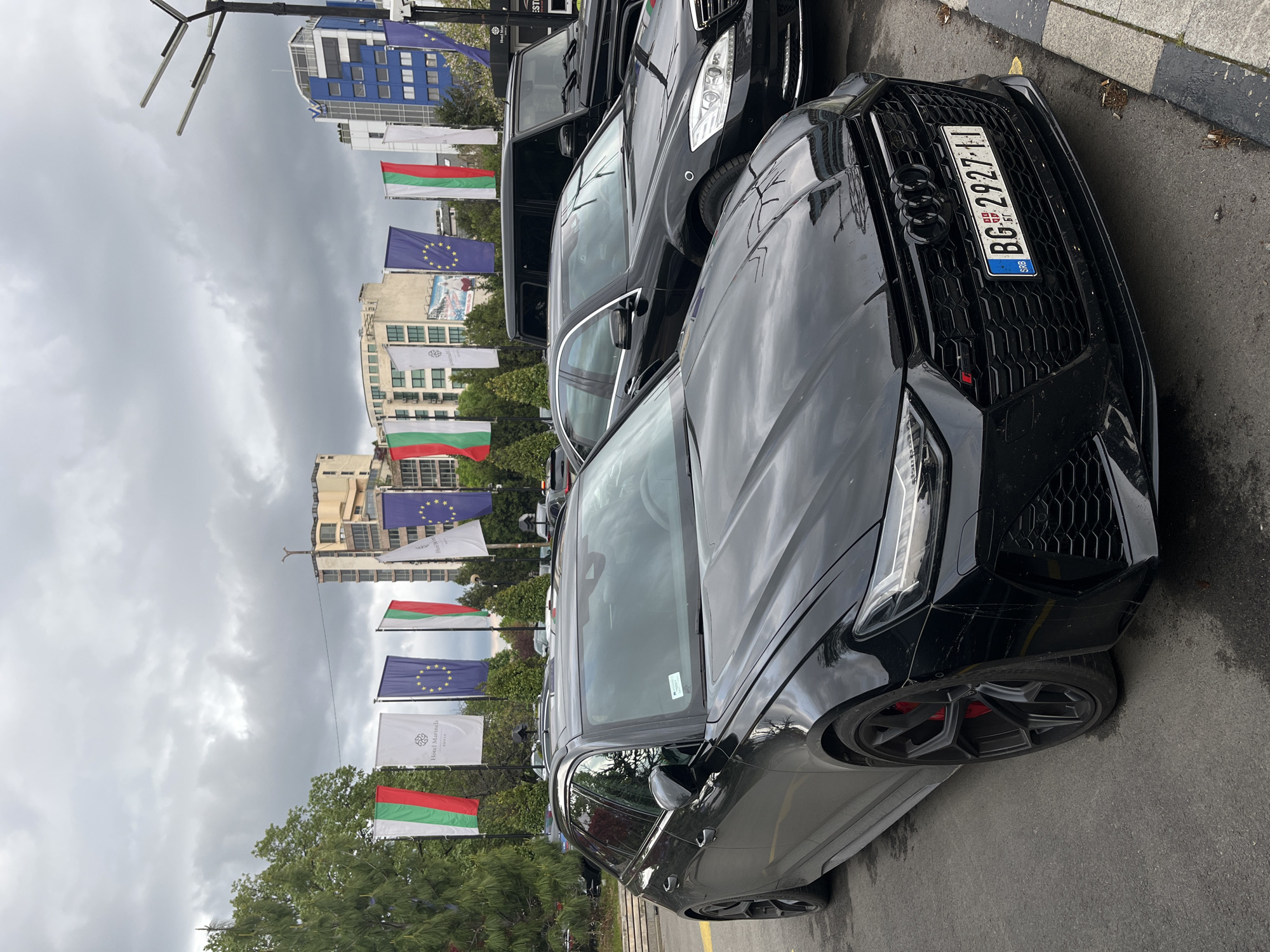 An outdoor, slightly rotated vertical shot captures a street scene dominated by parked luxury cars and a backdrop of flags and urban buildings under an overcast sky. In the immediate foreground, a black Audi RS model, likely an RS6 Avant, is prominently displayed. Its aggressive front grille with the Audi rings, modern LED headlights, and black multi-spoke wheels with red brake calipers are clearly visible. The car bears a Serbian license plate, "BG 2927-IJ," with "SRB" and "BG" (for Beograd/Belgrade) visible on the blue EU stripe.
Behind the Audi, several other dark-colored, high-end vehicles are parked, including what appears to be a Mercedes-Benz S-Class sedan and possibly a Mercedes G-Wagen, forming a line of prestigious cars.
The background features a row of flags flying from poles. These include multiple red, green, and white horizontal striped flags of Bulgaria, interspersed with blue flags bearing the circle of yellow stars of the European Union. Also visible are white banners, one distinctly displaying "Hotel Marinela Sofia" with a logo, indicating the location is near this hotel in Sofia, Bulgaria. Beyond the flags, various urban buildings with modern architecture can be seen, partially obscured by trees. The sky is uniformly cloudy, suggesting a daytime setting with diffused light. No people are visible, and the scene depicts parked vehicles in an urban environment.