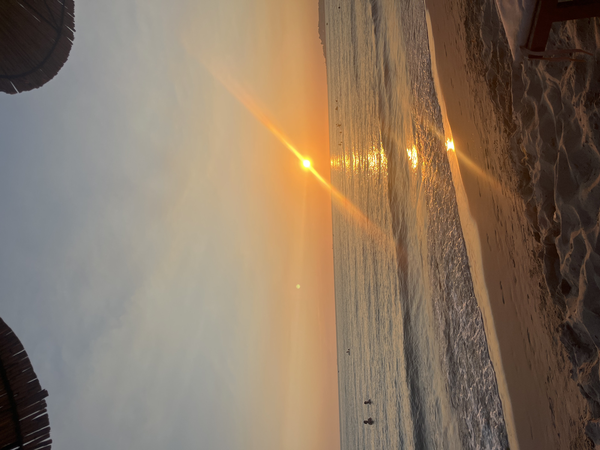 This image depicts a beach scene during sunset in the city of Bashkia Durrës, Albania. The sky is a gradient of orange and pale blue, with the sun low on the horizon, casting a bright, orange reflection on the water. A straw umbrella is visible in the upper left corner of the frame, and another in the upper right. The sea is calm, with gentle waves lapping at the sandy shore. Several people are visible in the water, some swimming and others on what appear to be paddleboards or small boats. The beach itself is sandy, with footprints visible on the wet sand near the water's edge. In the foreground, the sand is textured with dunes and shadows. In the far distance, a coastline with buildings can be seen. The overall impression is one of tranquility and natural beauty.