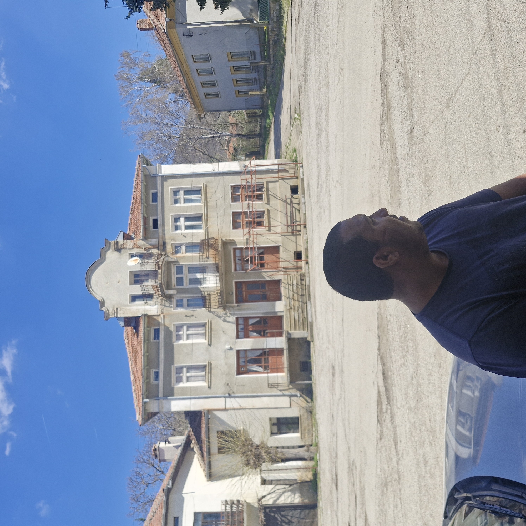 A daytime shot captures a man in a blue t-shirt standing next to a dark-colored car on a concrete surface. The scene is likely in Hirevo, Bulgaria, based on the information given. Behind the man and car, a multi-story building with a light-colored facade and red-tiled roof dominates the view. Scaffolding is visible on the building, suggesting ongoing construction or renovation. Adjacent to the main building are other structures, including what appear to be residential homes. A mostly clear, blue sky is visible above the buildings. A few bare trees are visible between the buildings. The man is looking upwards and towards the building. The photo appears to have been taken outdoors.
