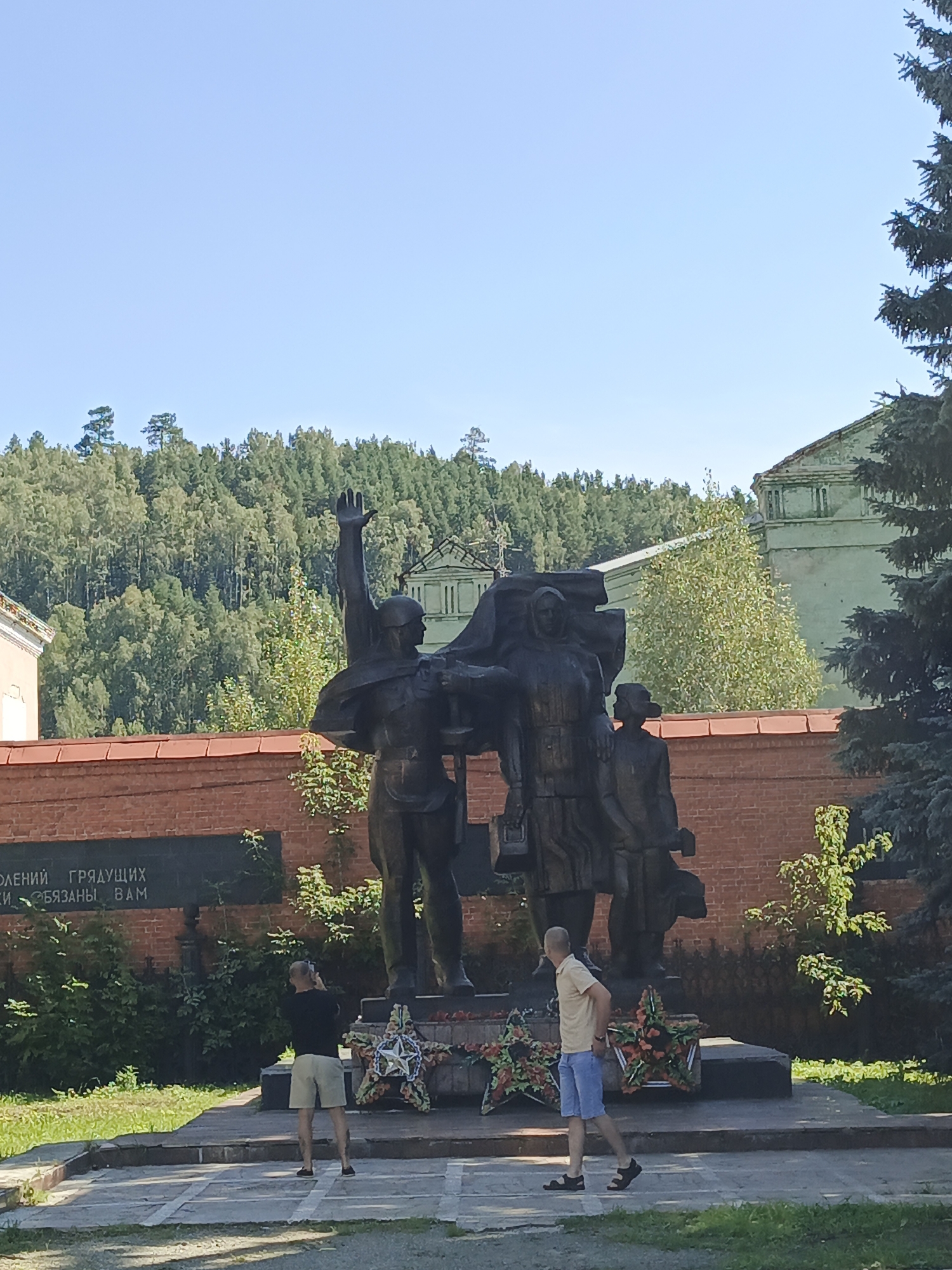 A bronze memorial monument, depicting three figures, is situated in a park-like setting in Zlatoust, Russia. The central figure, a soldier with his right arm raised, stands in front of two other figures, one of whom appears to be a woman holding a briefcase. The monument rests on a dark pedestal, adorned with two star-shaped wreaths at its base. Two men are present in the foreground. One man, wearing a black t-shirt and beige shorts, stands with his back to the camera, holding a mobile phone to his ear. The other man, in a yellow t-shirt and blue shorts, walks from right to left across the frame. Behind the monument is a low brick wall, and further beyond is a dense forest. The sky is clear and blue, and sunlight casts shadows on the ground. The monument is likely dedicated to the memory of soldiers and a testament to the legacy of those who fought in past conflicts.