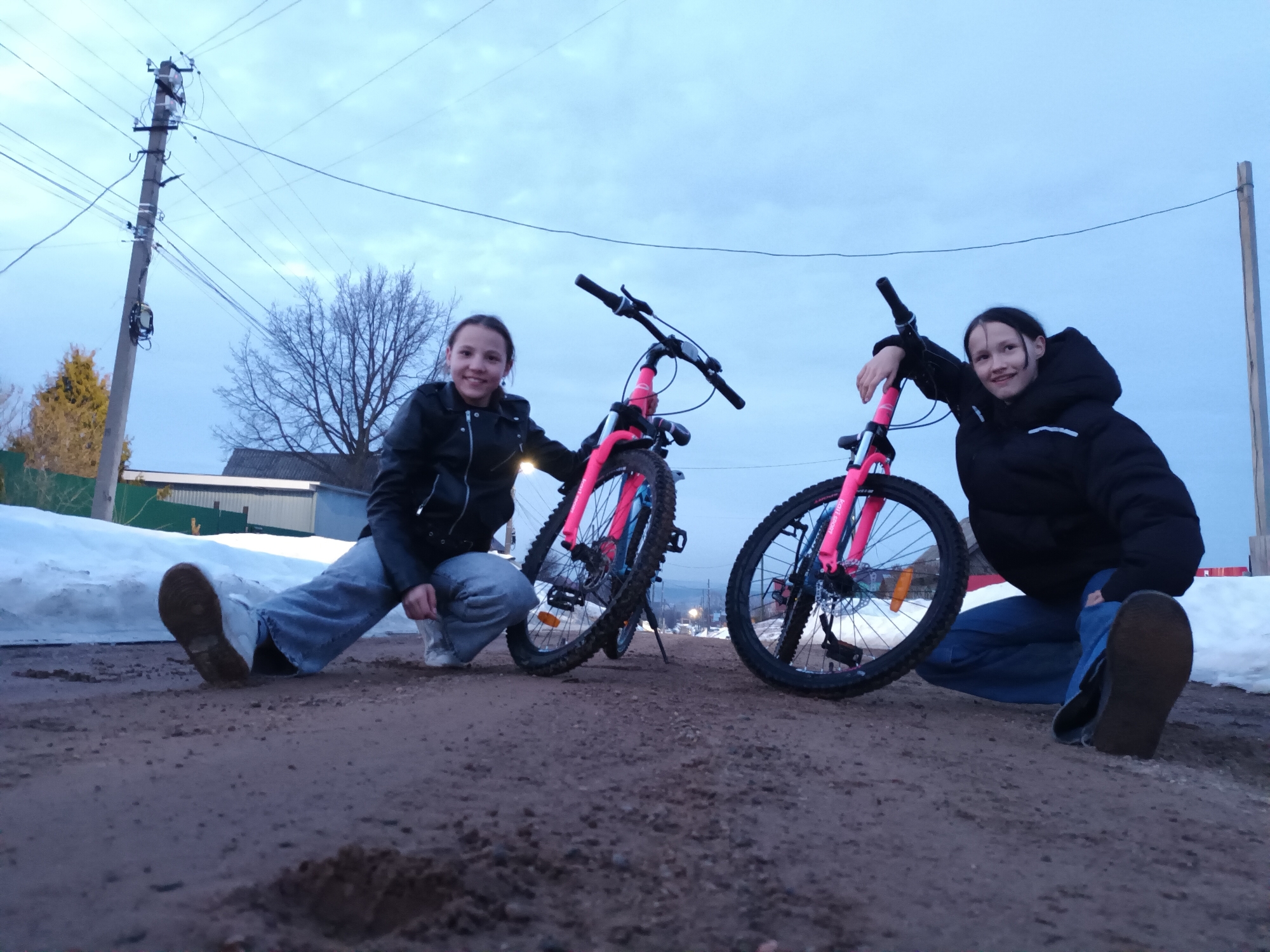 Two young females are depicted crouching on a dirt road in the city of Ольховка, Russia, posing with two identical pink-framed bicycles. Both subjects are smiling and looking directly at the camera. The female on the left wears a black leather-style jacket and light blue jeans, with her left hand resting on the handlebars of a pink mountain-bike style bicycle. The female on the right wears a black puffy jacket and light blue jeans, with her right hand resting on the handlebars of an identical bicycle.
The ground in the foreground is dark, uneven dirt or gravel. Significant snow banks are present along the sides of the road. In the background, a tall utility pole with multiple power lines is visible on the far left. A bare-branched deciduous tree is situated behind the subjects. Structures with dark roofs and fencing are partially visible further in the background. The sky is an overcast, light blue-grey.