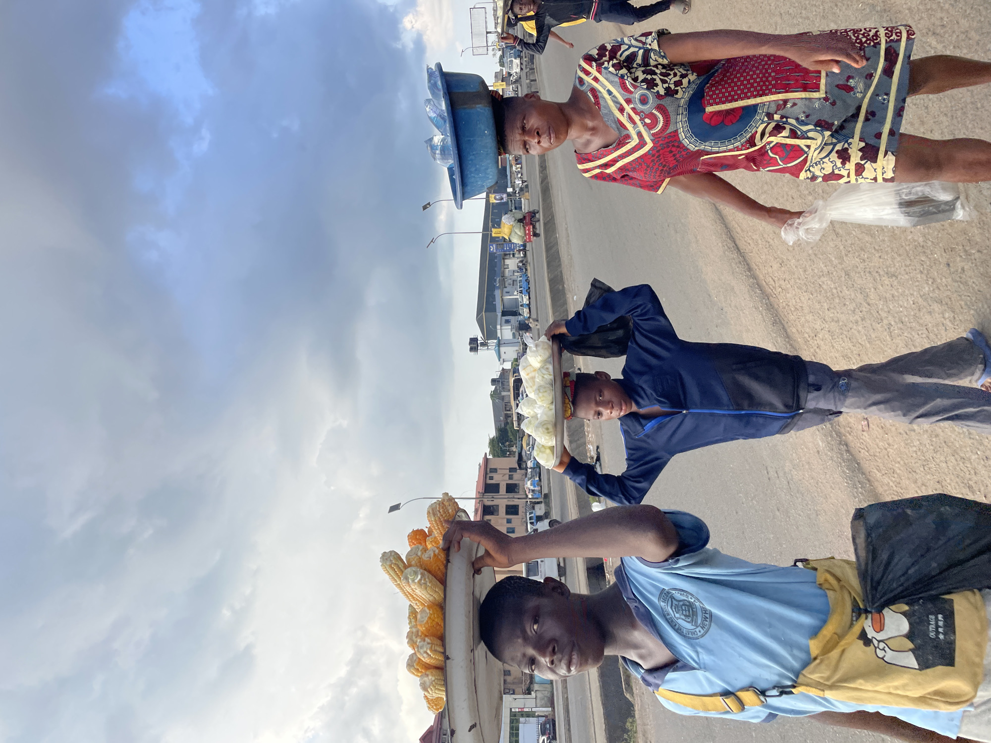 The image depicts three individuals on a street in Fegge, Nigeria, likely during the daytime given the clear sky with scattered clouds. The scene appears to be a busy road with vehicles and buildings visible in the background, suggesting an urban or semi-urban environment.
The primary focus is on the people and the goods they are carrying. In the foreground, a young boy in a blue t-shirt and yellow shorts is seen from the chest up, carrying a large platter of corn on his head. He has a black bag slung over his shoulder. To his left, another young person, wearing a dark blue jacket, is also carrying something on their head, which appears to be a white platter with what might be fruits or vegetables. Further back, a third person, dressed in a vibrantly patterned red, blue, and yellow outfit, is walking and carrying a large blue basin filled with bottles of water on their head, and holding a plastic bag with what seems to be an object inside.
The individuals appear to be street vendors, actively engaged in their trade, transporting and selling goods. The overall atmosphere seems to be that of daily commerce. There is no specific indication of the time of day beyond it being daylight, and the weather seems pleasant with a partly cloudy sky. The location cues are limited to what appears to be Nigerian street architecture and the context of Fegge, Nigeria. The expressions on the faces of the individuals are neutral or serious, common for people engaged in work. No discernible text is visible in the image.