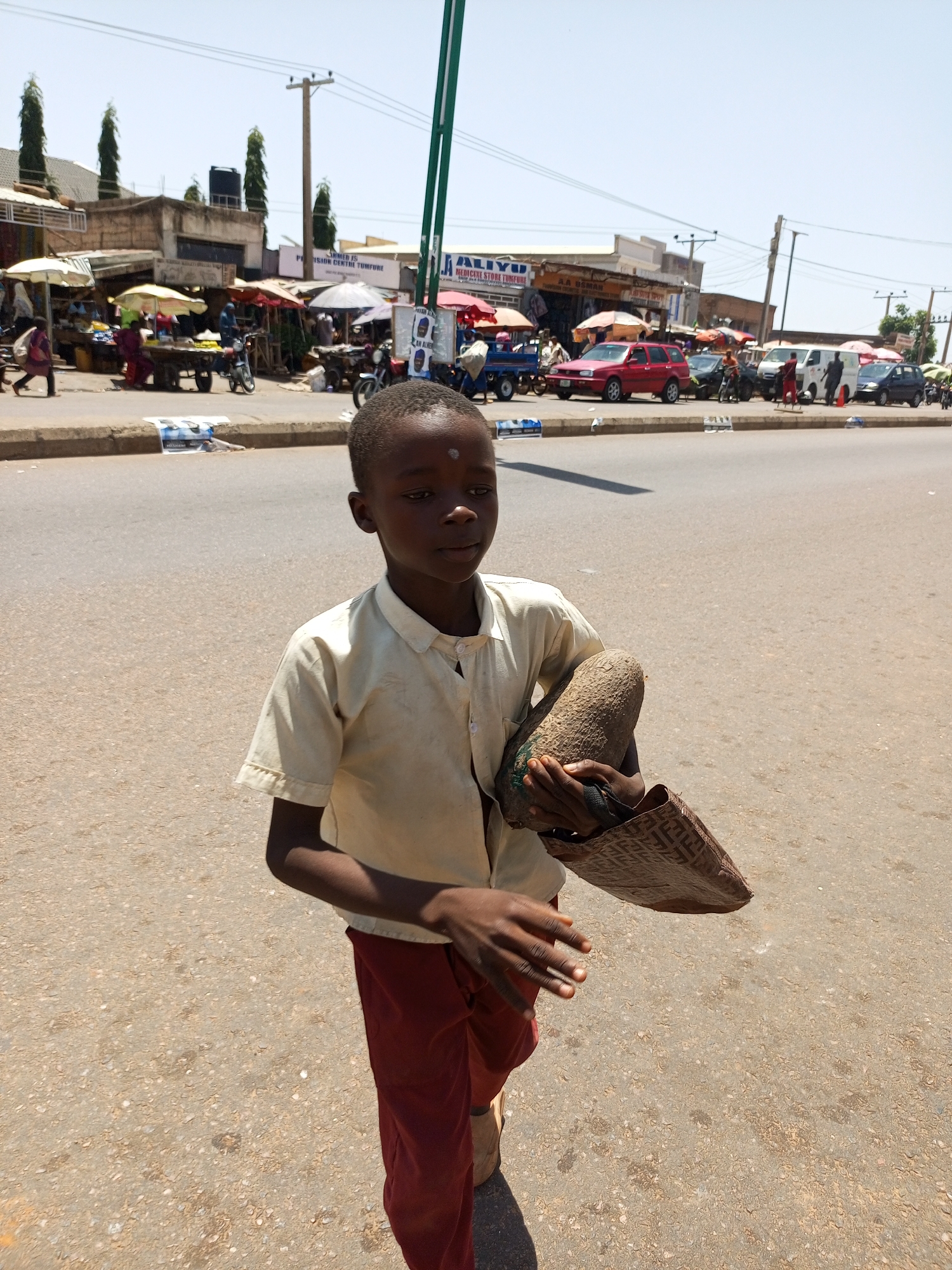 A young boy, likely of African descent, stands on a dusty road, holding what appears to be a large root vegetable and a woven bag. He is dressed in a light-colored, short-sleeved shirt and dark red trousers. The boy's expression is neutral, and he is looking slightly off to the right. His hands are visible, with one gripping the vegetable and the other holding the bag.
The background depicts a bustling street scene. Several market stalls with umbrellas are visible, suggesting a marketplace. Various people are moving around, and some are interacting with the stalls. A few motorcycles and cars are parked or driving on the road. Buildings line the street, some appearing to be shops with signage. The sky is clear and bright, indicating a sunny day, likely midday given the strong shadows cast. The overall setting appears to be a street market in a town or city.
Visible text includes "ALIVYO MEDICINE STORE TOMBURE" and "A.A USMAN" on signs of the shops in the background. The scene conveys a sense of everyday life in a busy market environment, with the boy being a central subject against the backdrop of commercial activity.