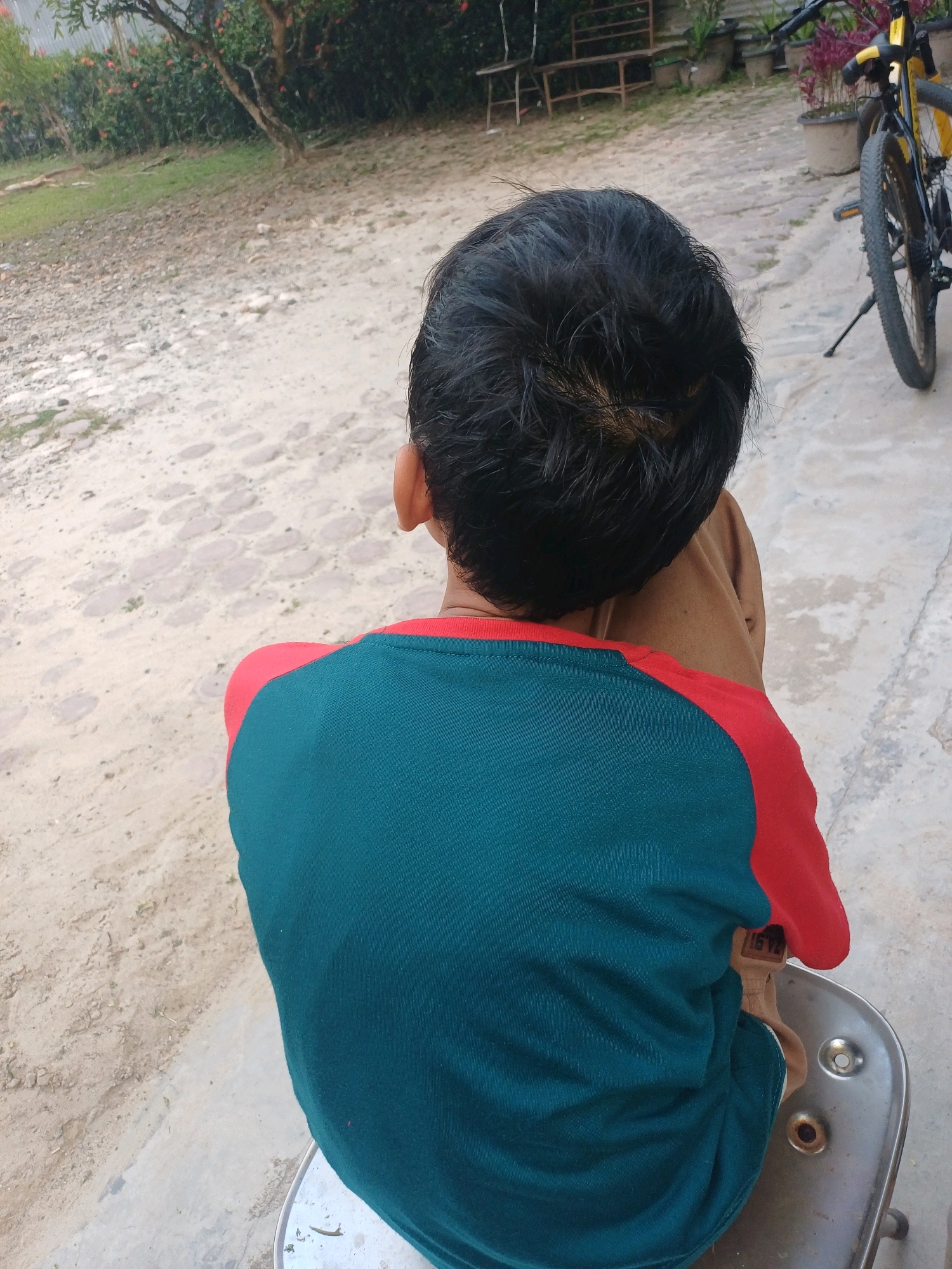 A boy, facing away from the camera, is sitting on a metallic chair. He has short, dark hair and is wearing a t-shirt with a teal body and red sleeves. His right arm is visible, and he is wearing brown pants. The boy is seated on a concrete surface. In the background, a bicycle with a yellow frame is parked. Further behind the bicycle, there are some potted plants. The area appears to be outdoors, with a sandy and paved ground. There is vegetation and trees visible in the distant background, and some makeshift wooden seating structures. The scene is likely in a residential or communal outdoor area within Dumai City, Indonesia.