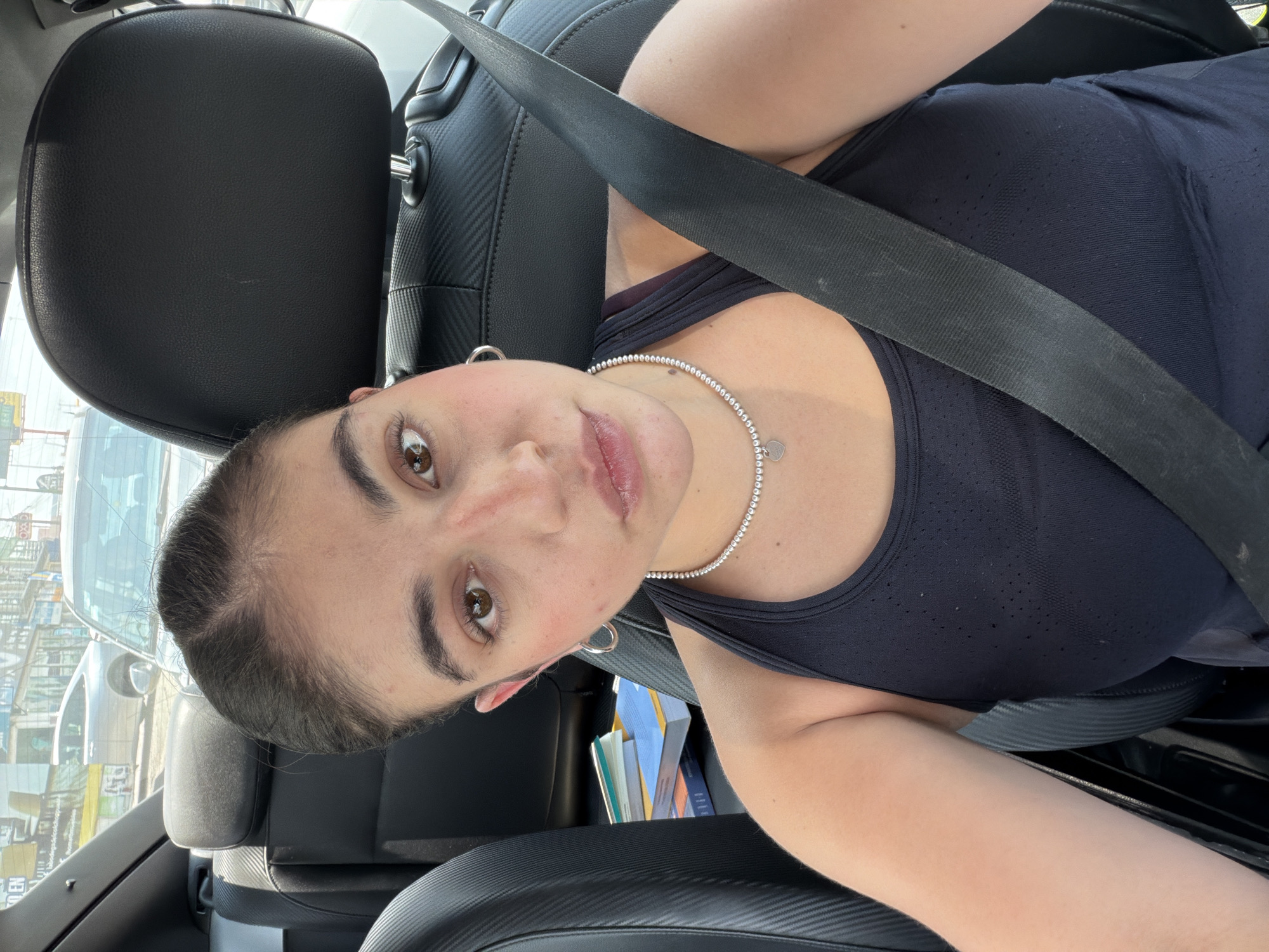 A young woman is captured in a selfie taken inside a car during what appears to be daytime. The image is rotated, presenting her leaning to the right from the viewer's perspective. She has a light to medium skin tone, brown eyes, and dark eyebrows, with her dark hair pulled back. She is wearing a dark, possibly navy or black, athletic-style tank top with subtle perforated detailing. Around her neck is a pearl-like necklace featuring a small, silver heart-shaped pendant. Small hoop earrings are visible on her ears.
She is seated in a car, evident from the black leather seats, a prominent black seatbelt crossing her torso, and car interior elements with a textured pattern. In the background, visible through the car window, are buildings, other vehicles, and some fragmented signs, suggesting an urban or semi-urban environment. A stack of books or papers is stored in a pocket or console next to her. The lighting is bright, indicating a sunny day. Her expression is neutral to slightly smiling. The location context provided is San Bernardino Tlaxcalancingo, Mexico. No discernible text is clearly visible on any signs or objects in the image.