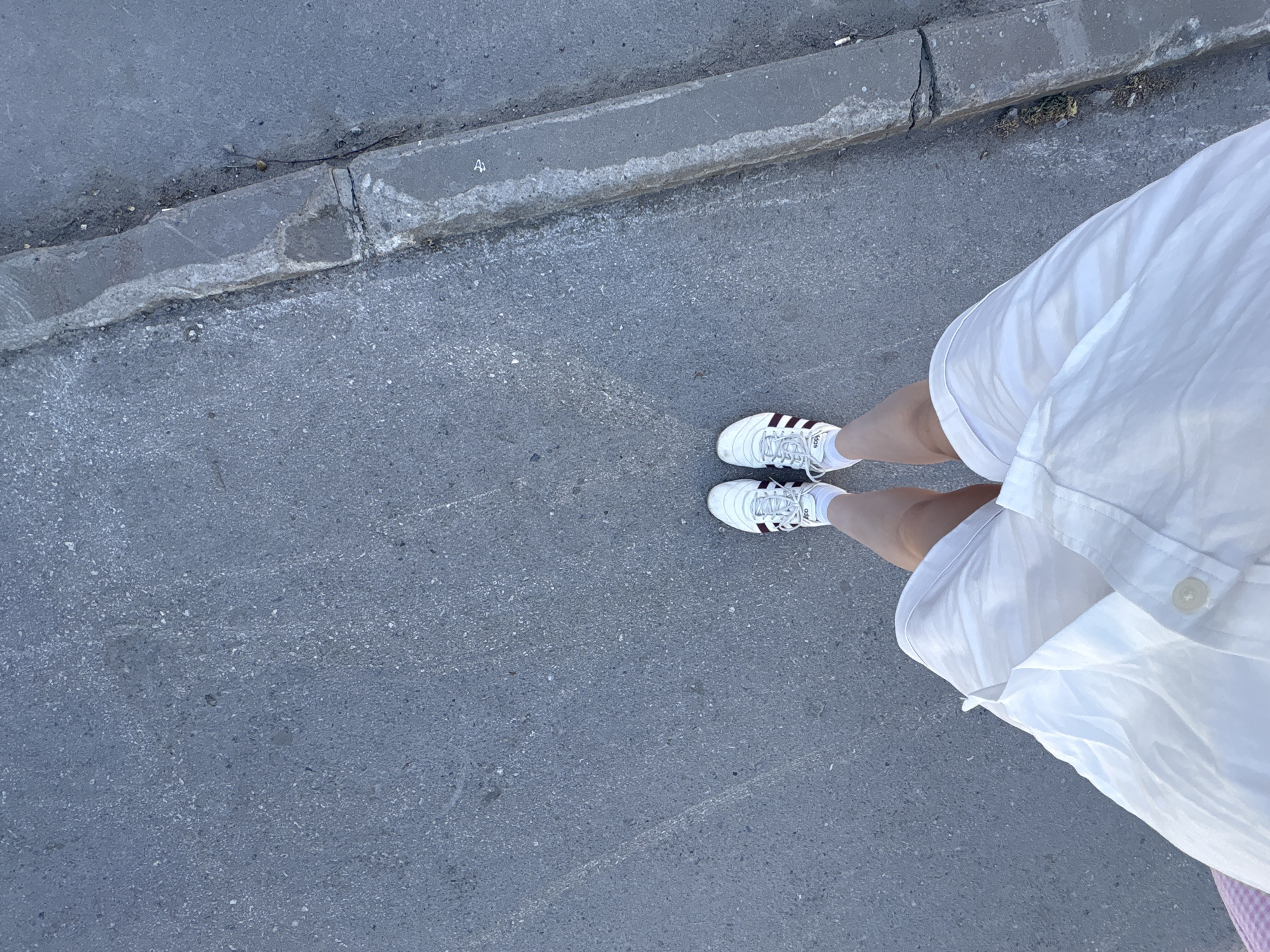 A high-angle shot depicts a person standing on an asphalt surface next to a concrete curb. The person is wearing white sneakers with maroon stripes, white socks, and white shorts. The asphalt surface appears to be a street or sidewalk. The location is Perm, Russia.