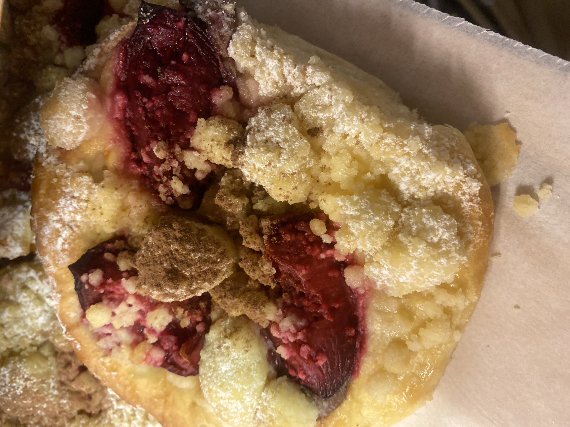 This media file features a close-up, overhead view of a single plum crumble pastry, presented on a piece of white paper. The pastry is round and appears to be baked to a golden brown. It is topped with large pieces of plum, some appearing halved or quartered, revealing their vibrant red-purple flesh and pits. Scattered over the pastry are generous clusters of crumble topping, which have a crumbly, golden texture. A dusting of powdered sugar lightly covers the crumble and the fruit. In some areas, the pastry and fruit are slightly glossy, suggesting it might be freshly baked or glazed. The overall impression is of a sweet, homemade baked good. The setting appears to be indoor, with the white paper suggesting it might be packaging or a serving liner. There is no visible text in the image. The lighting suggests an indoor environment, possibly with artificial light. The image does not contain any people or specific location cues beyond the given context of Warsaw, Poland, and the pastry itself is the sole object of focus.