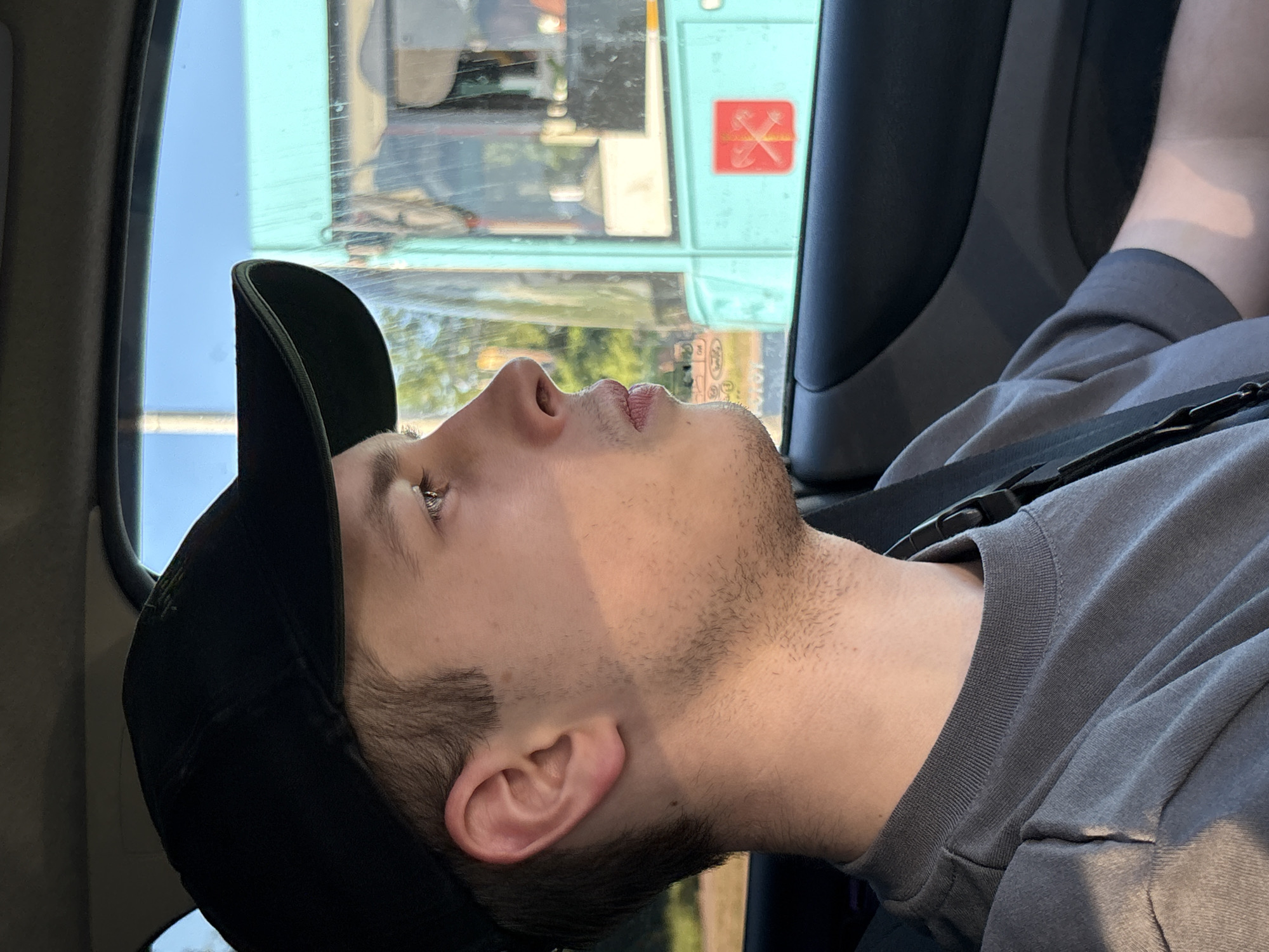 A young adult male occupies the passenger seat of a vehicle, his head turned in profile towards the right (viewer's left), gazing forward and slightly upward through the side window. He wears a black baseball cap with an indiscernible logo on the crown and a dark grey, short-sleeved, crew-neck t-shirt. His complexion is fair, with short brown hair visible under the cap and light stubble on his jawline and chin. Strong, low-angle sunlight illuminates the left side of his face and neck, creating a stark shadow line across his features. A dark strap, possibly a seatbelt or bag strap, crosses his chest diagonally.
The vehicle's interior features dark grey or black plastic trim on the door panel and window frame.
Adjacent to the vehicle, a large, light turquoise-colored public transport bus is visible through the car's window, its side windows reflecting the clear blue sky and surrounding green foliage. Prominently displayed on the bus exterior, below one of its windows, is a red rectangular emblem featuring two crossed, stylized golden/yellow axes or hammers. This specific emblem is historically associated with the Saint Petersburg Metro (Metropoliten) and is commonly seen on public transport within the city.
The bright blue sky and vibrant green foliage suggest fair weather during daylight hours. The scene is situated in the city of Saint Petersburg, Russia, specifically indicated by the distinctive public transport emblem on the adjacent bus.