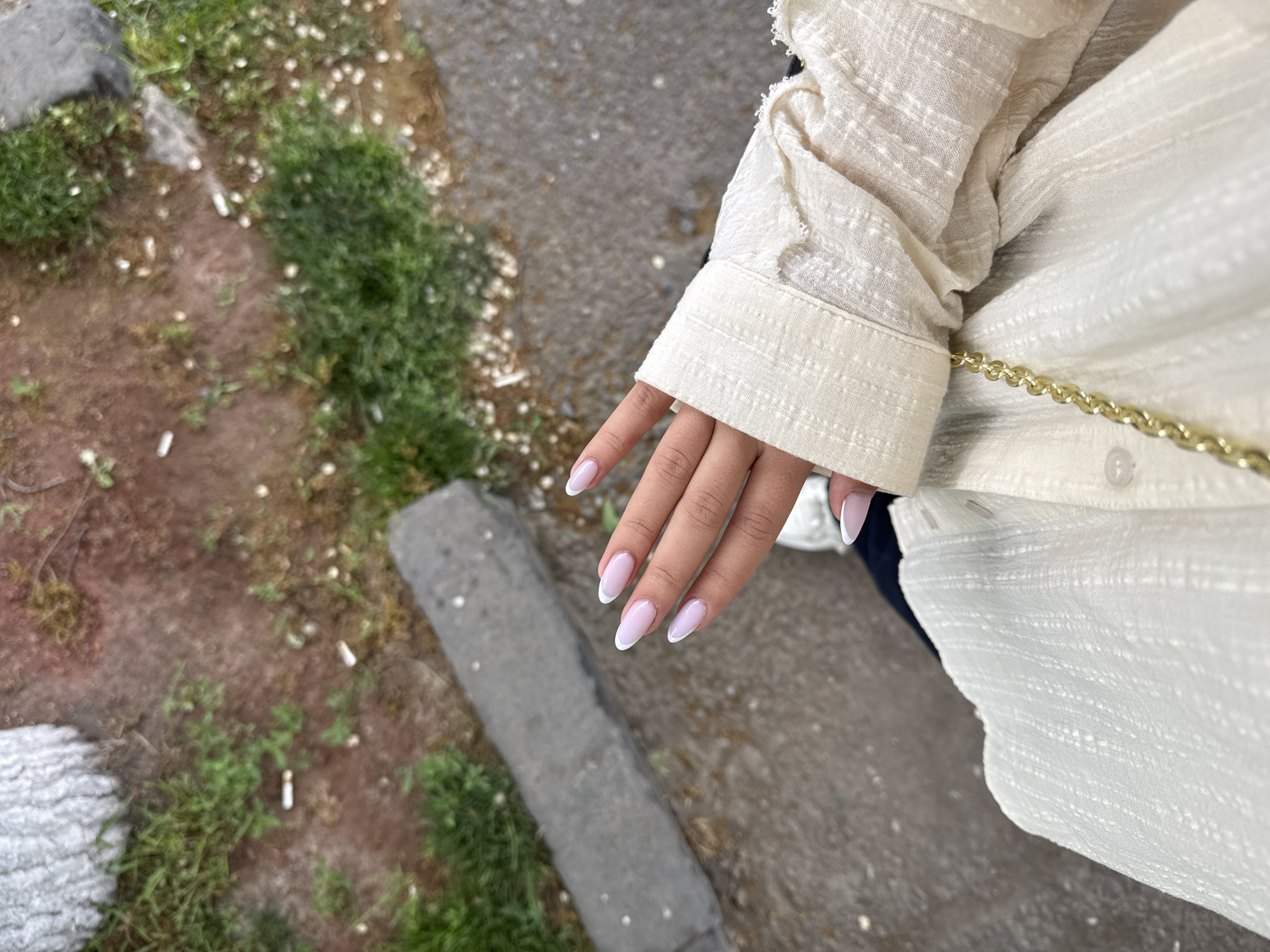A top-down shot shows a person standing outdoors, with a hand visible in the foreground. The person is wearing a light-colored, textured fabric shirt or jacket with a button and a gold chain detail on the garment. The hand has manicured nails with a white French tip design. The person is standing near a curb and a grassy area. The ground appears to be concrete.
The location is Yerevan, Armenia. The presence of a curb and grassy patch suggests an urban environment, possibly a street or park in the city.