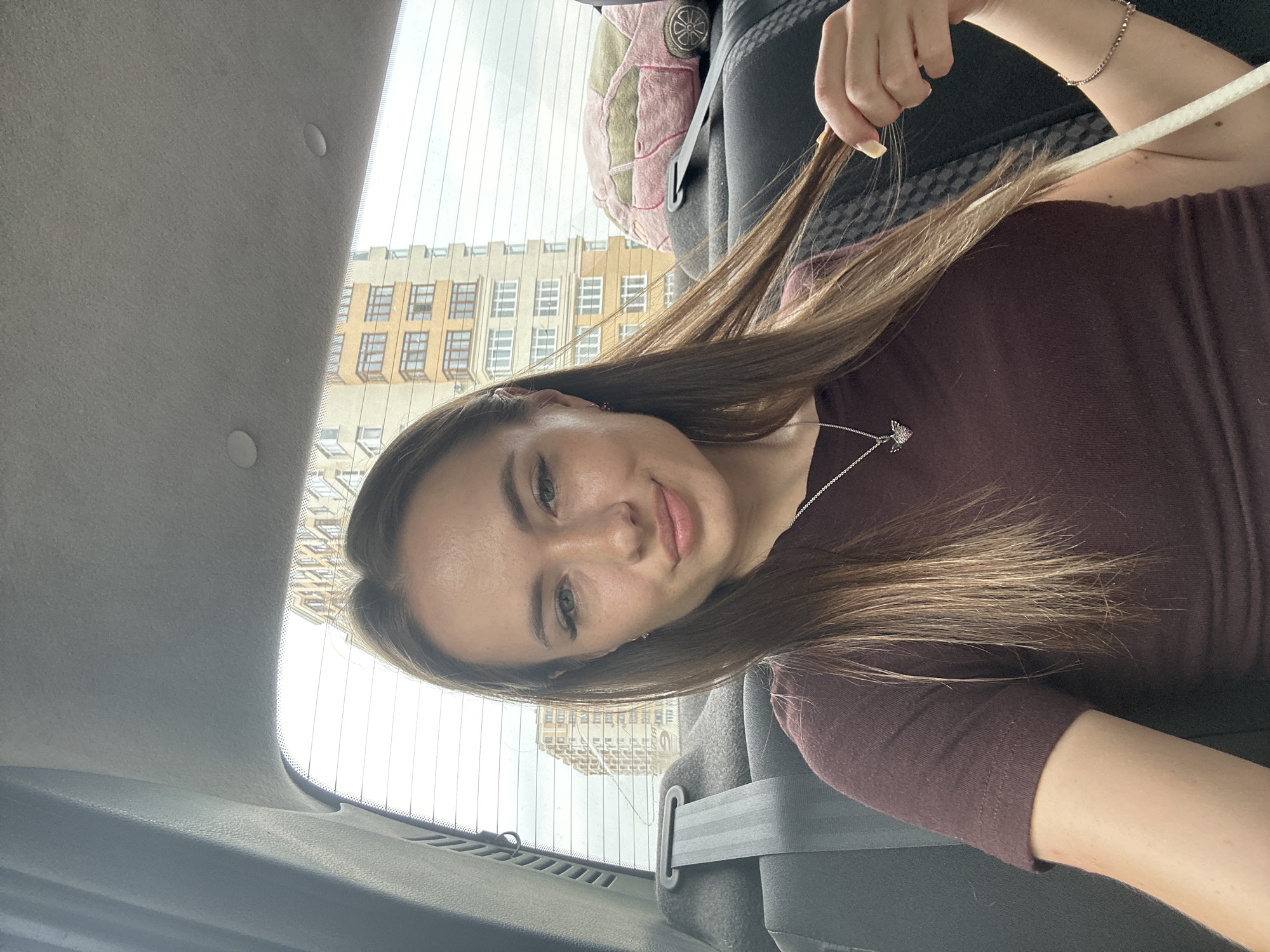 The image shows a young woman with long, light brown hair, wearing a dark maroon t-shirt and a silver necklace with a heart pendant. She is sitting in the back seat of a car, with a seatbelt visible. She is holding a strand of her hair and looking at the camera with a slight smile. Through the car's rear window, two tall buildings with multiple windows are visible, suggesting an urban environment. A pink plush toy in the shape of a car is also visible on the seat behind her.