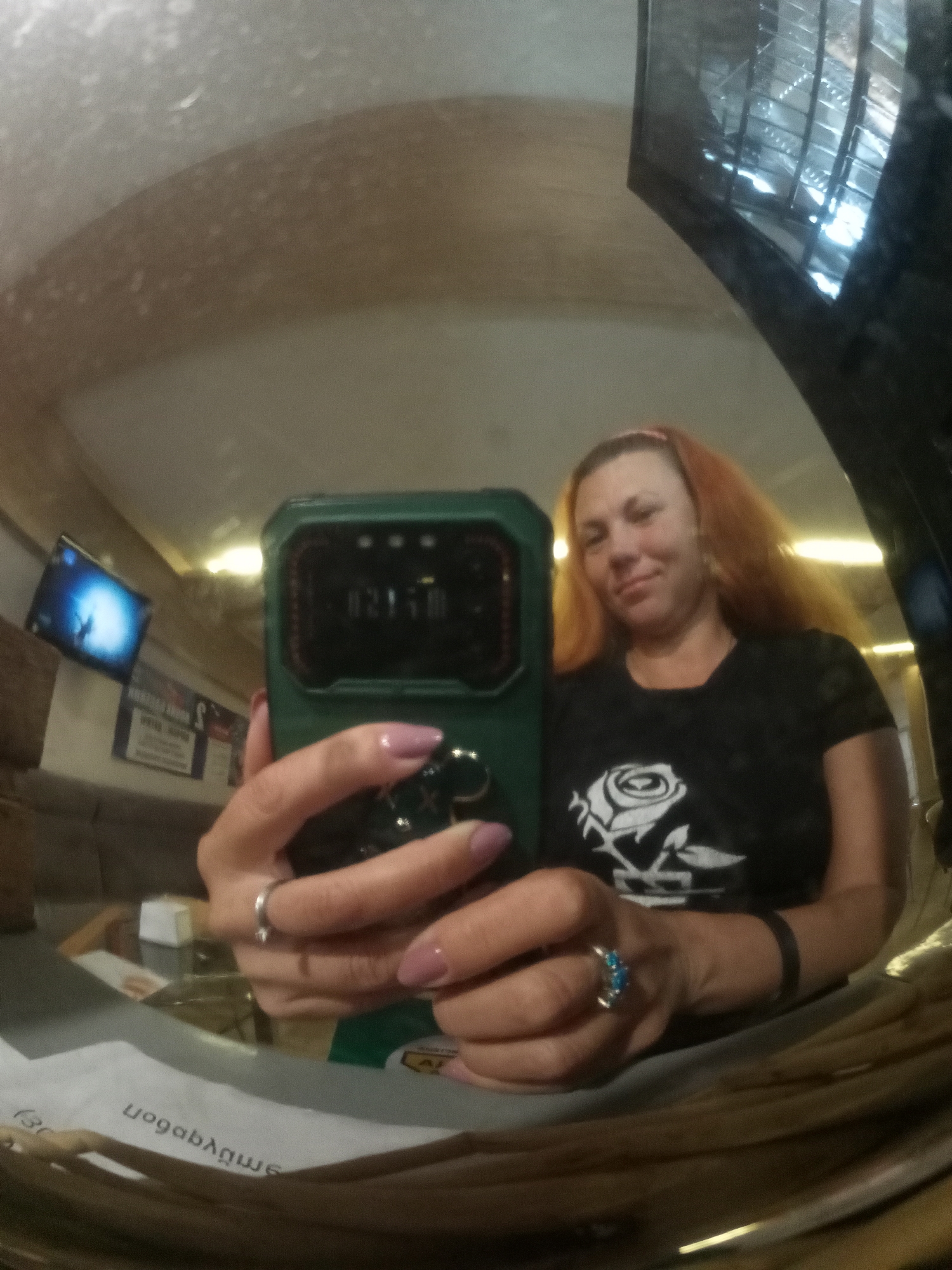 A woman with red hair is taking a selfie in a wide-angle mirror. She is wearing a black t-shirt with a white rose graphic. Her right hand is holding a green phone, and her left hand is resting on her thigh. She has several rings on her fingers, including a silver band with blue stones on her left ring finger and a thin silver band on her left index finger. The reflection in the mirror shows a television screen in the background. The setting appears to be indoors, possibly a bar or restaurant, with overhead lighting and what looks like a metal grate ceiling. There is text visible on a white surface in the lower part of the mirror's reflection that appears to be reversed, suggesting it's a reflection.
