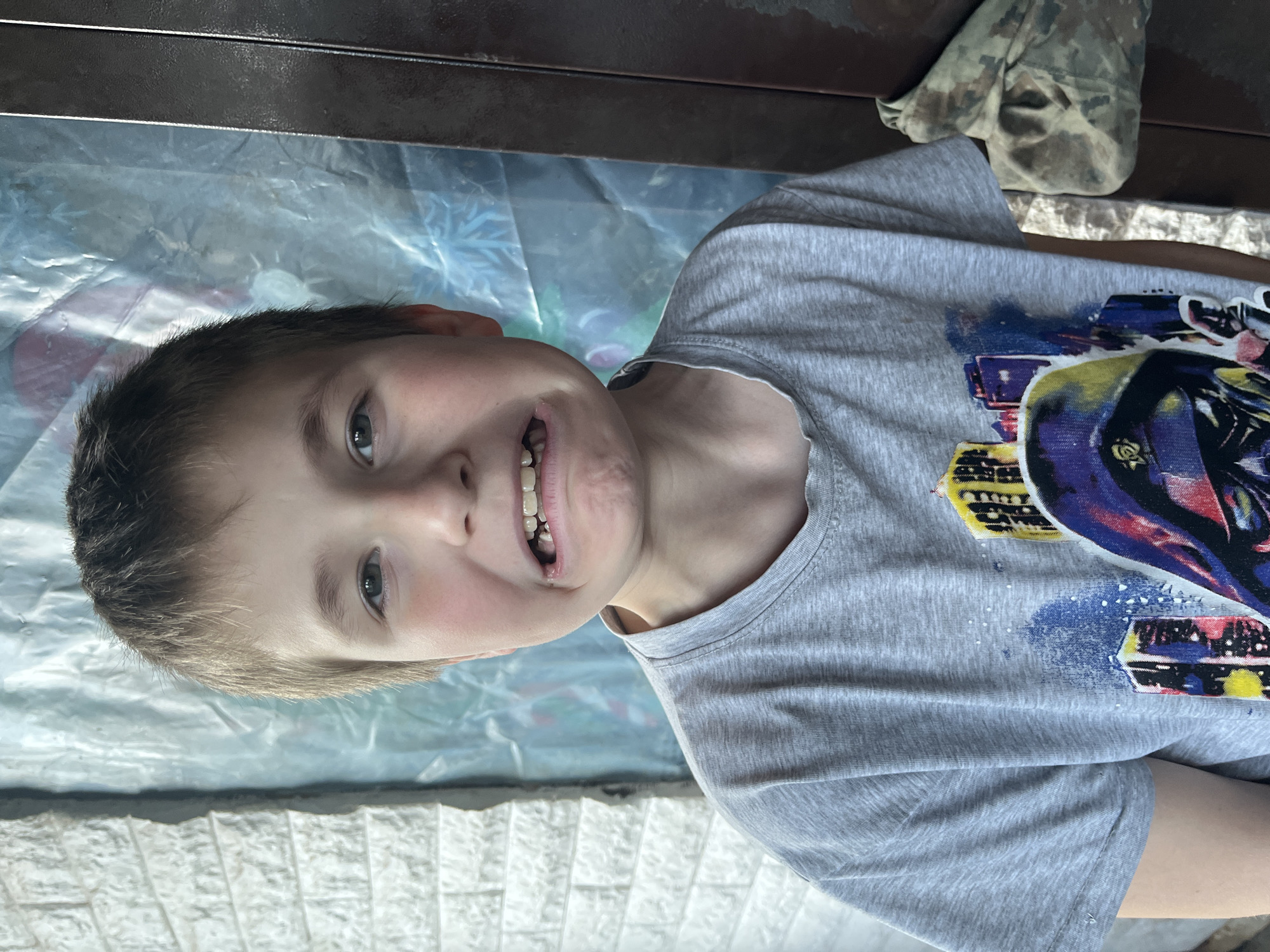 A young boy with short brown hair is wearing a gray t-shirt with a colorful design. He is smiling with his upper teeth visible. The boy is positioned on what appears to be a clear plastic surface or tablecloth, with a dark-colored wooden or metal surface visible above. To the upper right of the boy is a cloth object with a camouflage pattern. The photograph appears to be taken outdoors, based on the natural lighting. No clear indication of the location, Vesyoloye, Russia is visible.