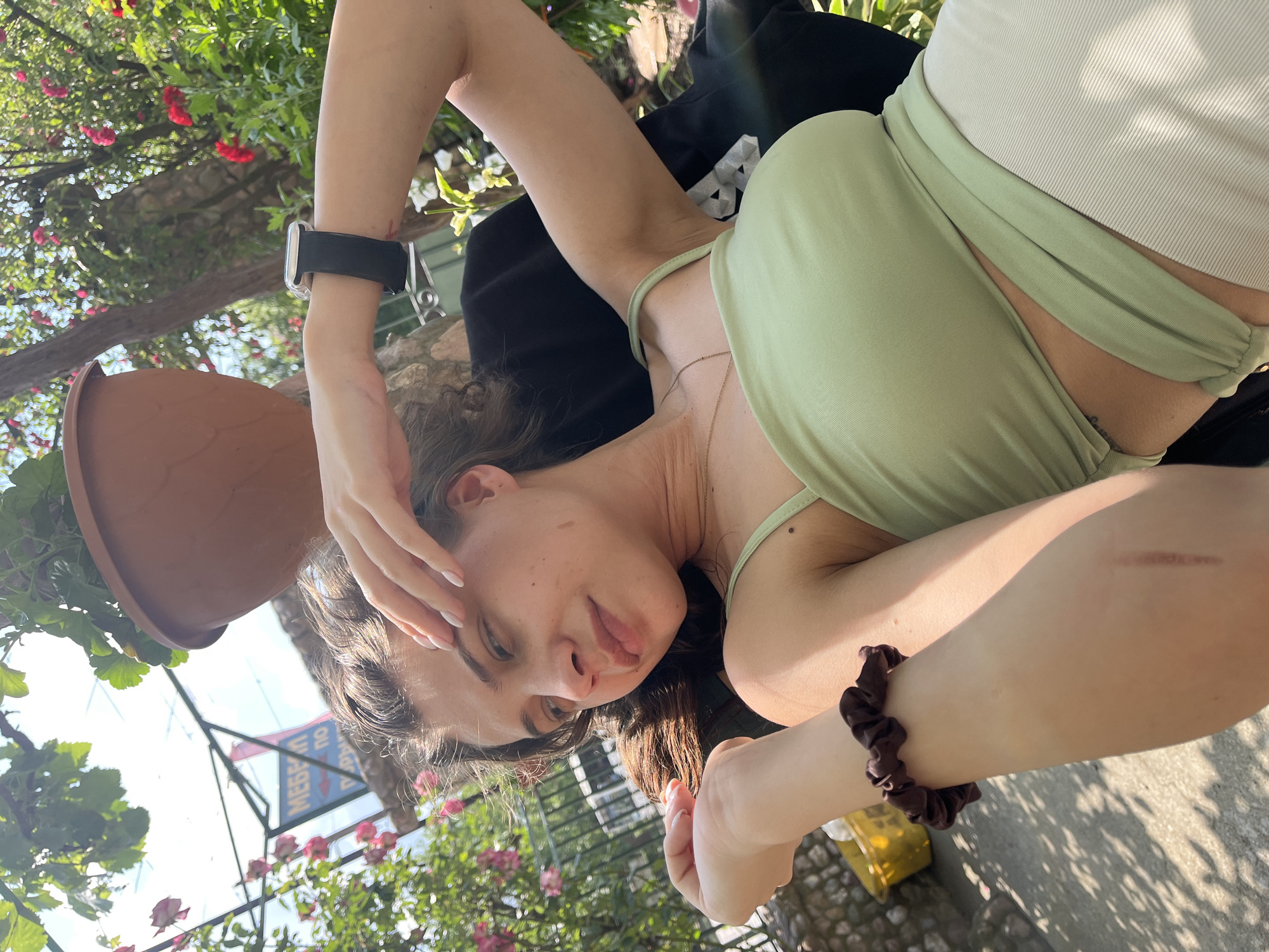 A young woman with long, dark, wavy hair is captured in a relaxed, reclining pose, looking towards the camera. She appears to be lying on her back or side, with her head resting on her left arm, which is bent. Her right hand is gently touching her hair or forehead. She is wearing a light green, halter-neck style top and light-colored bottoms, possibly a skirt or shorts, made of a ribbed fabric.
On her left wrist, she wears a black smartwatch with a clear or silver-toned frame. A delicate gold-colored chain necklace is visible around her neck. On her right wrist, she has a dark brown scrunchie, and a healed scar is noticeable on her right forearm. Her nails are manicured with a light, natural polish. A small mole is visible on her left upper arm/shoulder area.
The setting is outdoors, characterized by abundant lush green foliage and flowering plants with red, pink, and some white blooms, suggesting a garden or patio. A terracotta potted plant is prominent in the foreground. The lighting indicates a bright, sunny day, casting distinct shadows on the paved ground. In the background, hints of structures, possibly a building or awning, are visible. There's a clear container with a yellow-green beverage on a surface to her right.
Visible text in the background is in Cyrillic script, reading "МЕБЕЛ" (Mebel, meaning furniture) and possibly parts of other words like "ПОДАРЫ", indicating a location in a Slavic or Russian-speaking country. The overall mood is casual and serene.