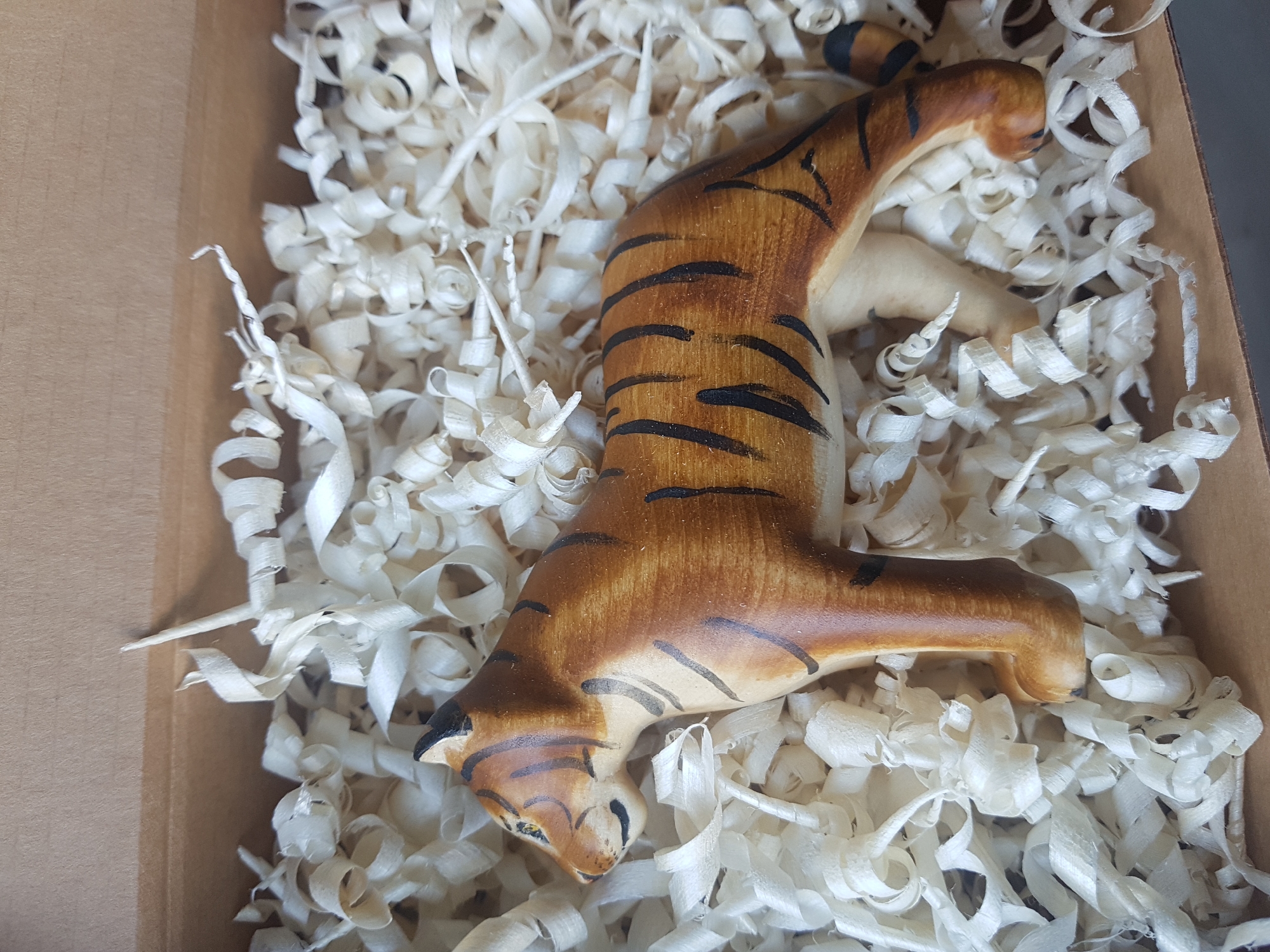 A wooden tiger figurine lies on a bed of white wood shavings inside a cardboard box. The tiger is painted with brown and black stripes, and its head is turned to the left. The box is brown and appears to be open on the side. The scene has a well-lit environment. There is no specific indication of the location being in Saint Petersburg, Russia.