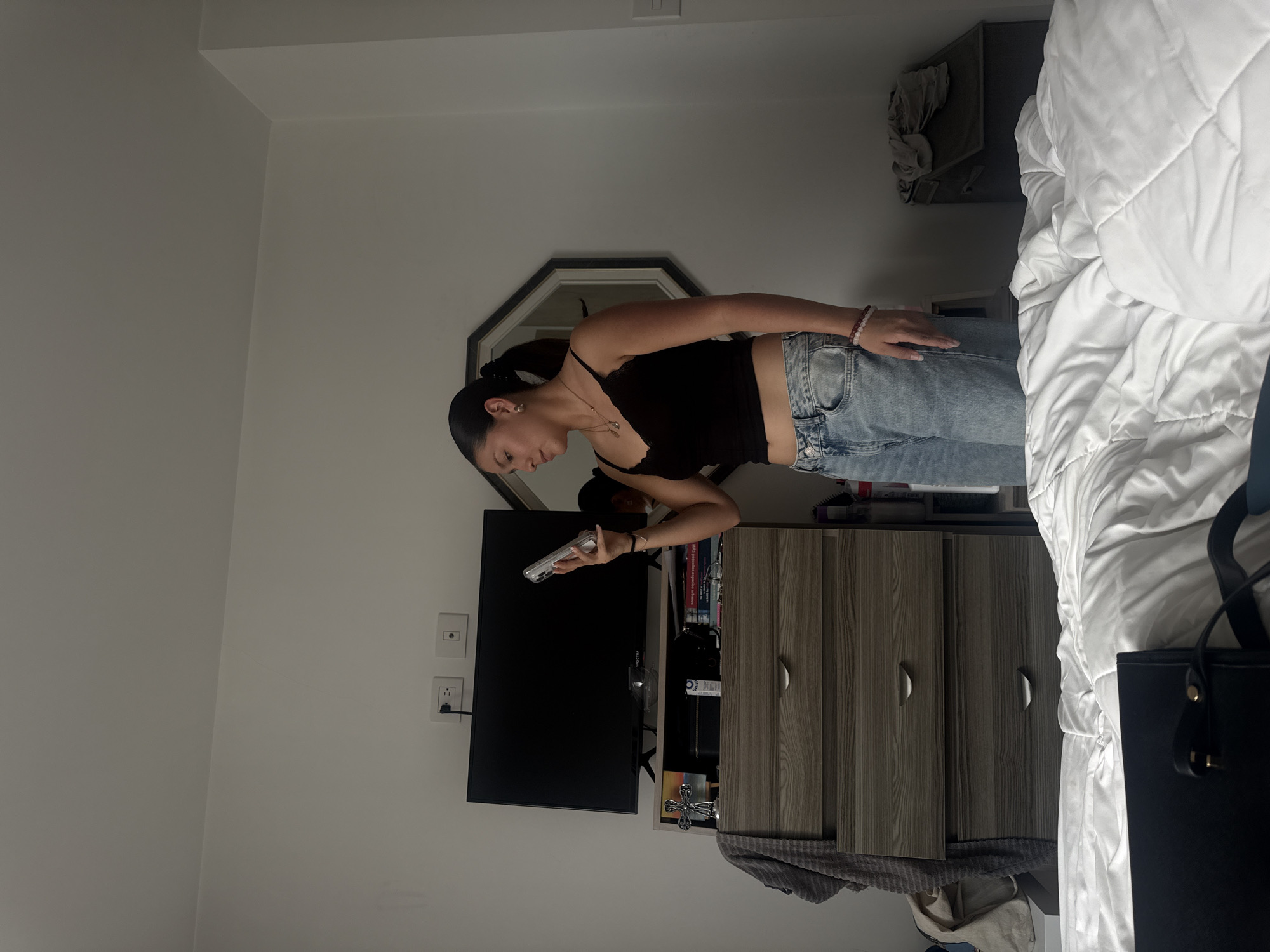 A woman with long dark hair pulled back in a ponytail is standing in a bedroom, looking down at a phone she is holding. She is wearing a black top and light blue jeans. Behind her is a television mounted on a wall above a dresser. A mirror hangs above the television. A partially visible bed with white bedding is to the right. The room appears to be in a private residence, but the location in San Francisco Ocotlán (Ocotlán), Mexico is not visually confirmable.