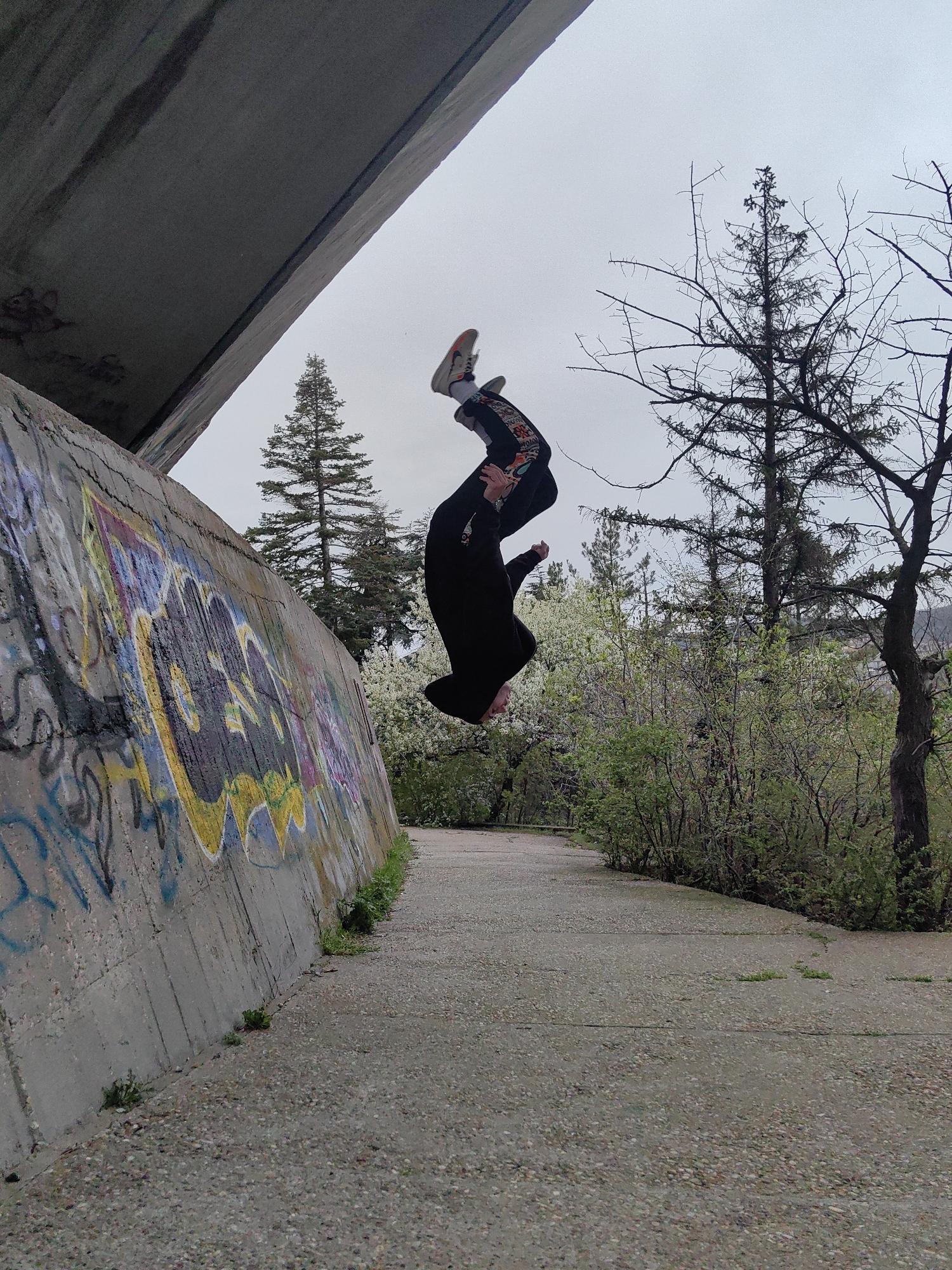 A person is captured mid-air performing a backflip, with their body nearly vertical and head pointing downwards. The setting appears to be an outdoor concrete walkway bordered by vegetation on the right and a graffiti-covered concrete structure on the left, possibly a bridge support. Coniferous and deciduous trees are visible in the background. This location is likely in the city of Varna, Bulgaria.