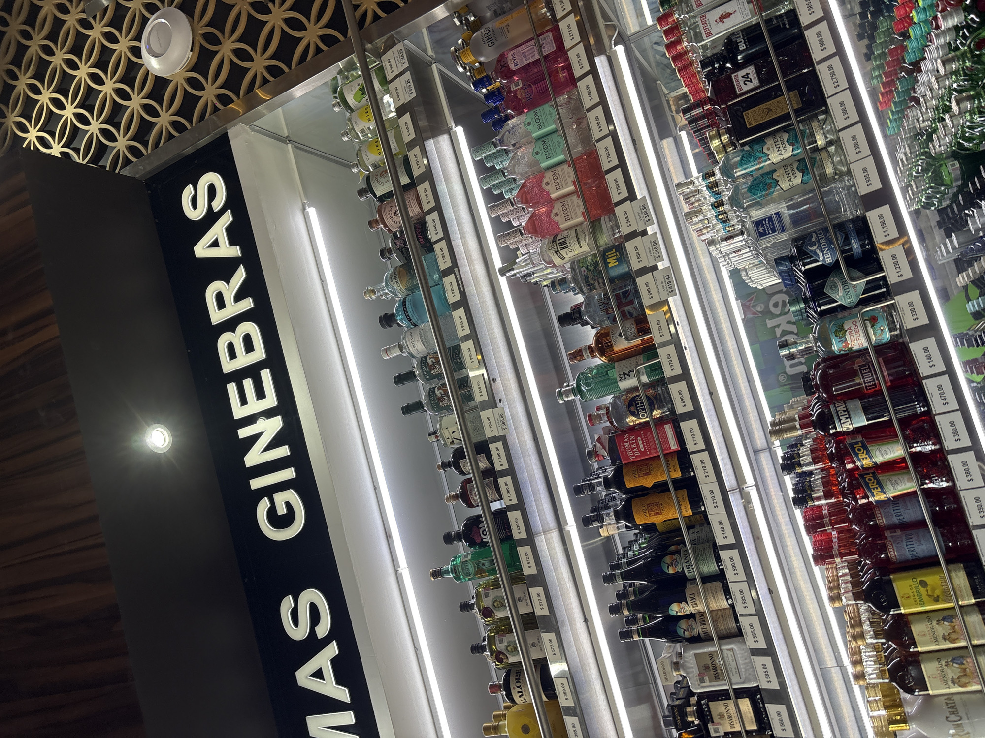 The image shows a retail display of gin bottles in San Bernardino Tlaxcalancingo, Mexico. A sign reading "MAS GINEBRAS" (More Gins) is visible on the left. The gin bottles are arranged on shelves with price tags. Different brands are visible including "BLOOM," "HENDRICK'S," and "BEEFEATER 24."