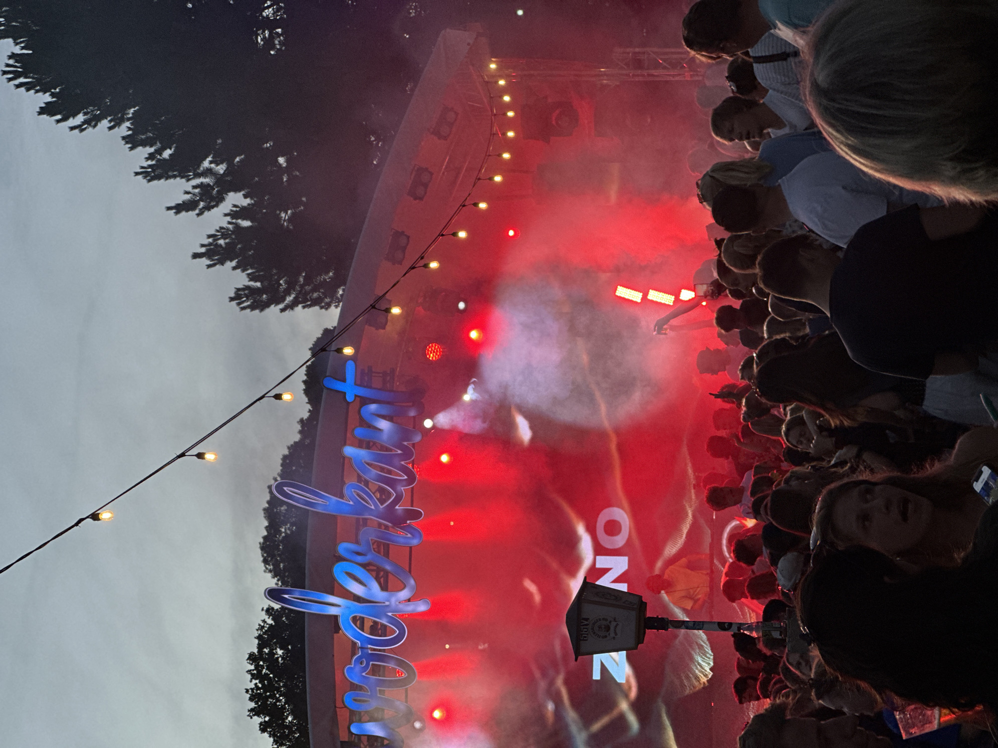 This media file captures a lively outdoor event, likely a concert or festival, with a crowd of people gathered to enjoy the festivities. The scene is illuminated by vibrant red stage lighting and smoke, creating a dramatic and energetic atmosphere. A large, illuminated blue sign with cursive lettering is prominently displayed, though the full text is not entirely legible. The words "superheart" are visible. Other objects present include string lights with yellow bulbs, a speaker or sound equipment, and part of a stage structure. The time of day appears to be evening or night, given the artificial lighting and the darkening sky visible at the top of the image. Trees with dark foliage frame the scene, suggesting an outdoor venue possibly in a park or open space. The crowd is a mix of individuals, with some looking towards the stage, others conversing, and a general sense of engagement and excitement. The overall mood is festive and dynamic, indicative of a well-attended public gathering in Kiel, Germany.