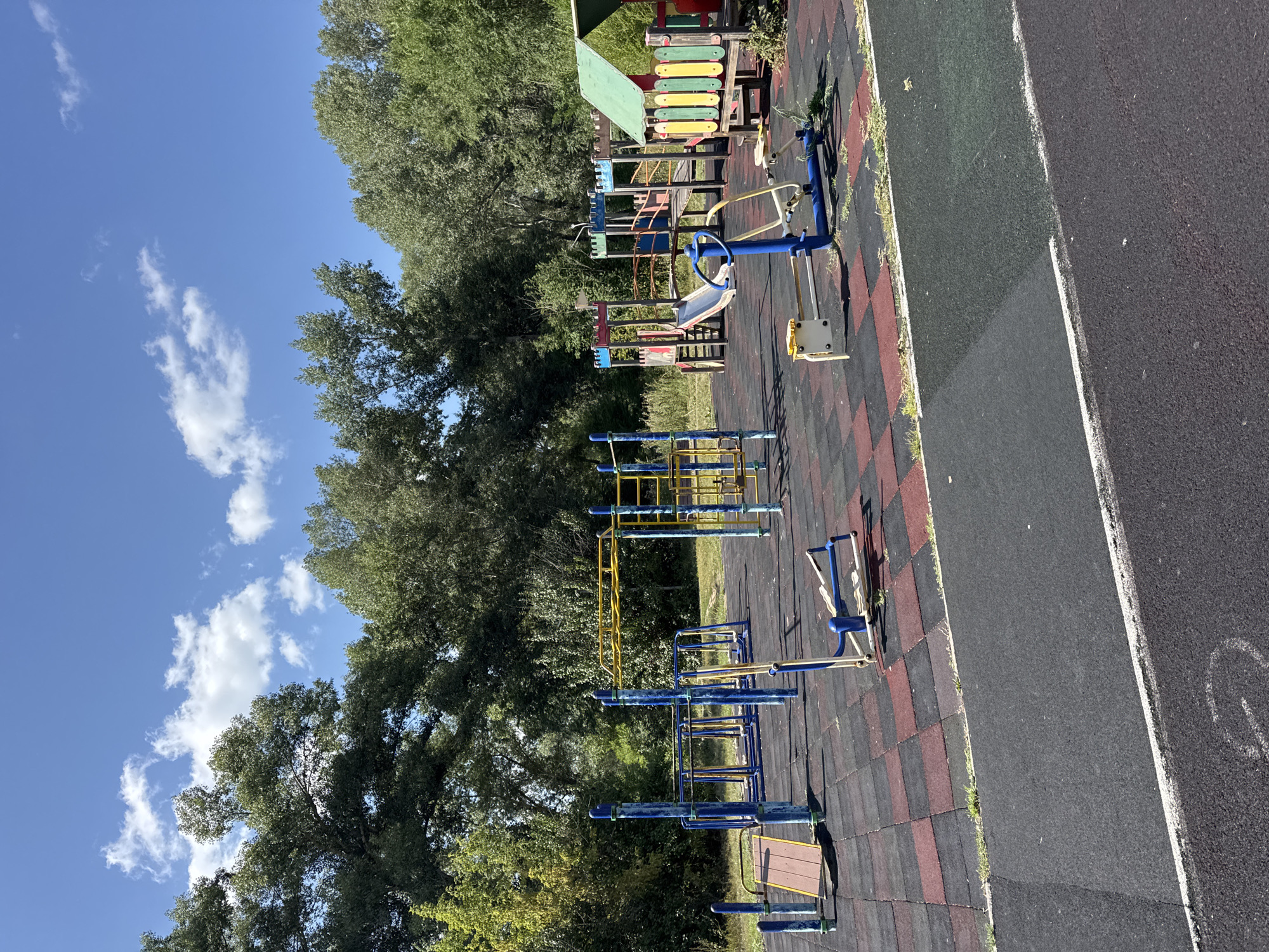 A public playground is depicted on a sunny day, surrounded by lush green trees. The ground is covered with a black and red rubberized surface, interspersed with black asphalt paths. Various playground equipment is visible, including a yellow monkey bar structure, blue and yellow climbing frames, a slide, and individual exercise machines. In the background, a dense line of trees forms a natural backdrop against a clear blue sky with scattered white clouds. The scene appears to be located in a park or recreational area. The image does not show any people, actions, or interactions. There are no signs or indications that the location is in the city of Oskemen, Kazakhstan.